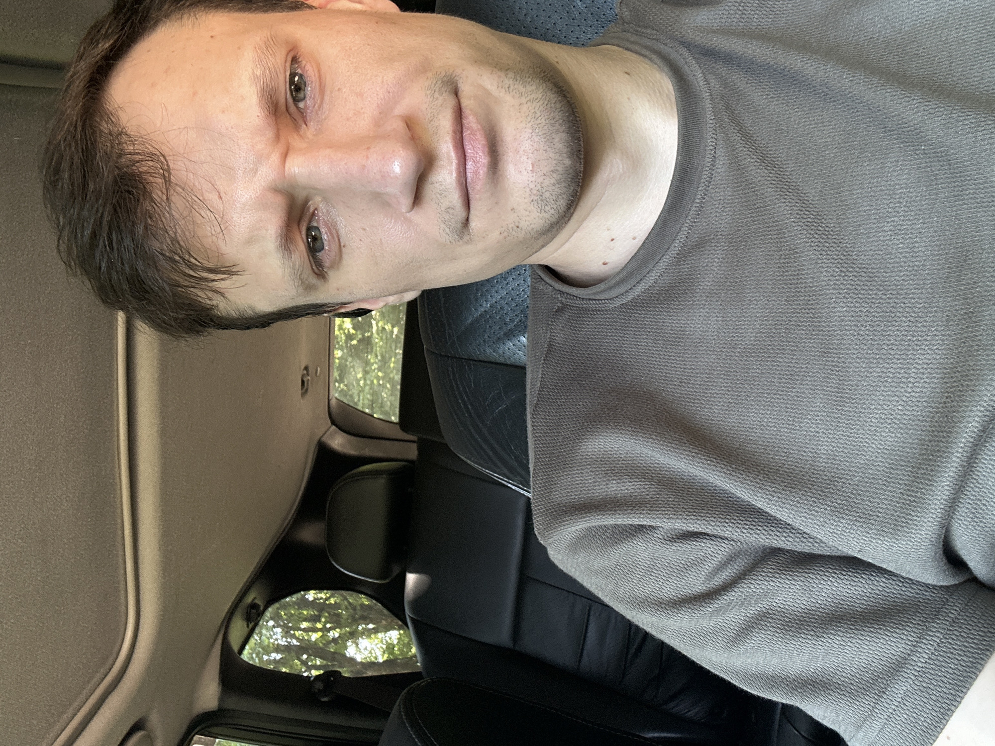 In Shatava, Ukraine, a man with short brown hair and a slight stubble is captured in a close-up selfie from inside a vehicle. He wears a brown, textured t-shirt. His expression is one of slight annoyance or concern, with furrowed brows and narrowed eyes. The interior of the car is visible, featuring black leather seats and a sunroof. Outside the car, sunlight filters through green trees.