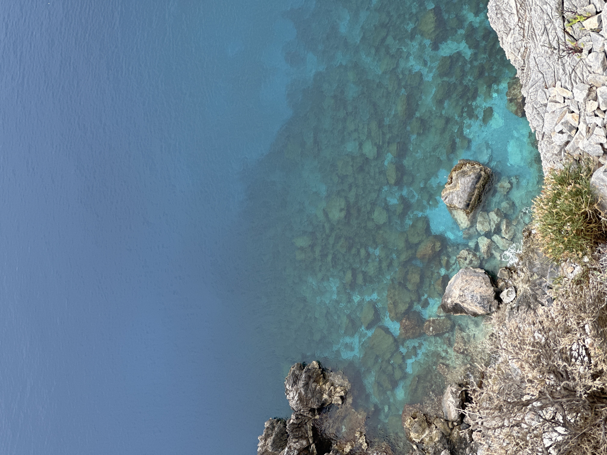 The image presents an elevated view of a tranquil coastal scene in Datça, Turkey. The majority of the frame, spanning the left and central areas, is dominated by the calm, blue waters of the sea.
The water on the far left appears a uniform, deep, and opaque blue, suggesting significant depth. Moving towards the right, the water becomes progressively clearer and lighter, transitioning into a vibrant turquoise. Through this translucent water, the rocky seabed is clearly visible, revealing numerous formations, boulders, and pebbles in shades of brown, grey, and green.
On the right side of the image, a rugged, light-colored rocky coastline descends into the sea. This shoreline is characterized by jagged rock formations and scattered stones. Several large, dark rocks are positioned along the water's edge, some partially submerged and appearing to have marine growth.
Sparse, hardy vegetation, including small green bushes and drier, gnarled shrubs, clings to the rocky terrain on the right. The scene is bathed in bright, natural daylight, indicative of a clear, sunny day. No human activity or visible text is present. The overall atmosphere is one of natural beauty and serenity.