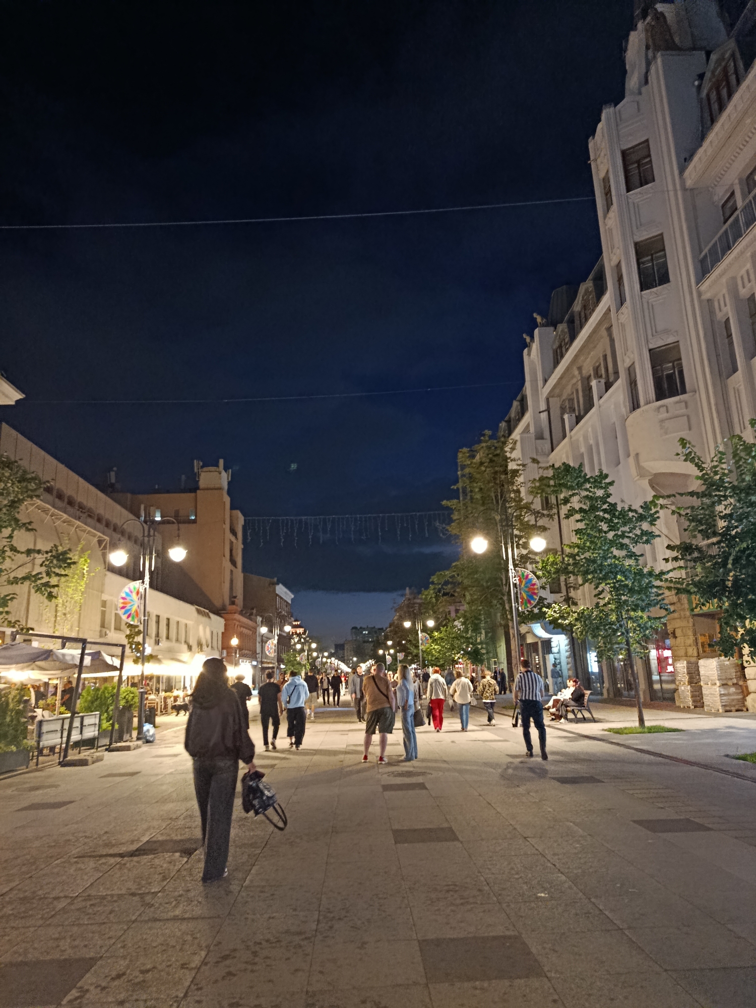 The image depicts a nighttime scene on a pedestrian street in Saratov, Russia, likely during warmer months given the attire of the individuals. The street is lined with buildings on both sides, illuminated by streetlights and lights from businesses. On the left, there are cafes with outdoor seating under awnings. On the right, the buildings are taller and more ornate, featuring balconies and architectural details. A string of decorative lights hangs across the street in the distance.
Several people are walking along the street. In the foreground, a woman with dark hair, wearing dark clothing, walks away from the camera, carrying a backpack. Further down the street, various individuals and small groups are seen strolling. Some are engaged in conversation, while others appear to be simply enjoying the evening. The street is paved with large, rectangular stones. Trees are planted along the sides of the street, with some foliage visible. The sky is dark, indicating it is night.