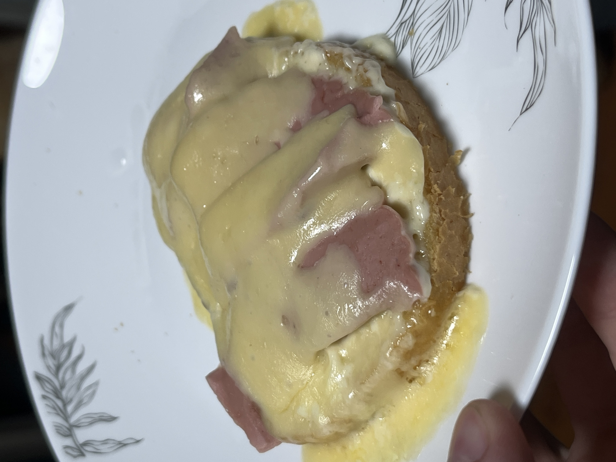 A close-up, eye-level shot shows a single piece of toast topped with ham and melted cheese on a white plate. The toast is golden brown and sits slightly askew. Slices of pink ham are visible beneath the creamy, pale yellow cheese, which is melted and slightly browned in places. The cheese has a glossy, gooey texture and drips over the sides of the toast. The plate has a subtle leaf-like design around the rim. A human finger is visible in the lower left corner of the frame, holding the plate. The background is dark and out of focus, suggesting an indoor setting. The image appears to be taken in Nizhny Tagil, Russia, likely in a home kitchen or dining area.