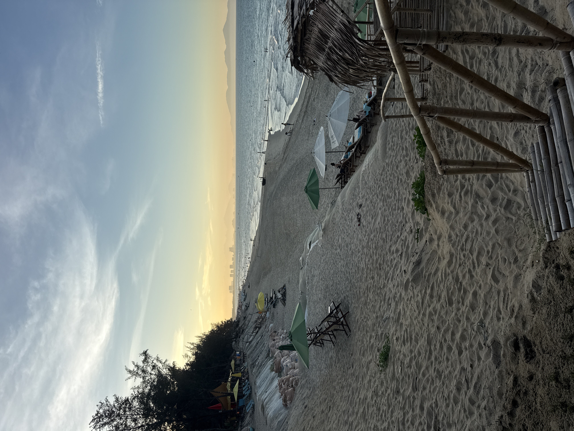 The image shows a beach at an indeterminate location, possibly at sunset or sunrise, as evidenced by the golden sky. The beach has a variety of beach umbrellas in green, white, and yellow, positioned along the shore. Reclining chairs are set up under some of these umbrellas, suggesting a leisure area.
The beach is sandy, with the foreground showing detailed sand texture and minor vegetation. The ocean meets the beach in the midground, with small waves breaking. In the background, a coastline or mountain range is visible, and a silhouette of buildings can be seen.
A bamboo structure, possibly part of a building or beach infrastructure, is present on the right side of the image, partially obstructing the view. People can be seen walking on the beach and relaxing in the chairs.