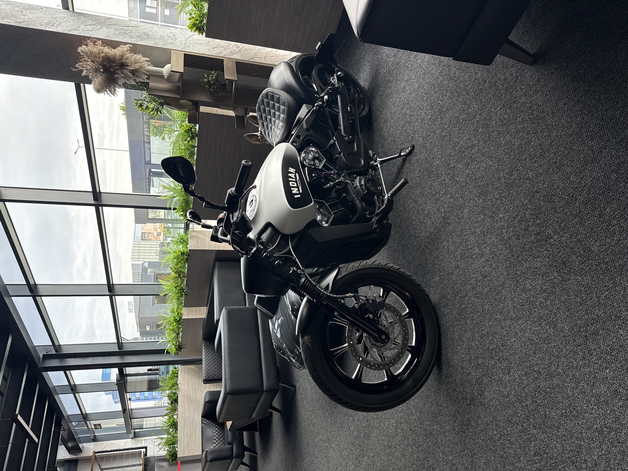 The image depicts an Indian Scout motorcycle indoors. The motorcycle is predominantly black and white, with the brand name "INDIAN" visible on the gas tank. The bike is parked on gray carpet next to modern seating consisting of black chairs and sofas.
In the background, a glass wall reveals an urban landscape with buildings and foliage. Based on information visible through the windows and the architectural style, the location is likely in the city of Plovdiv, Bulgaria.