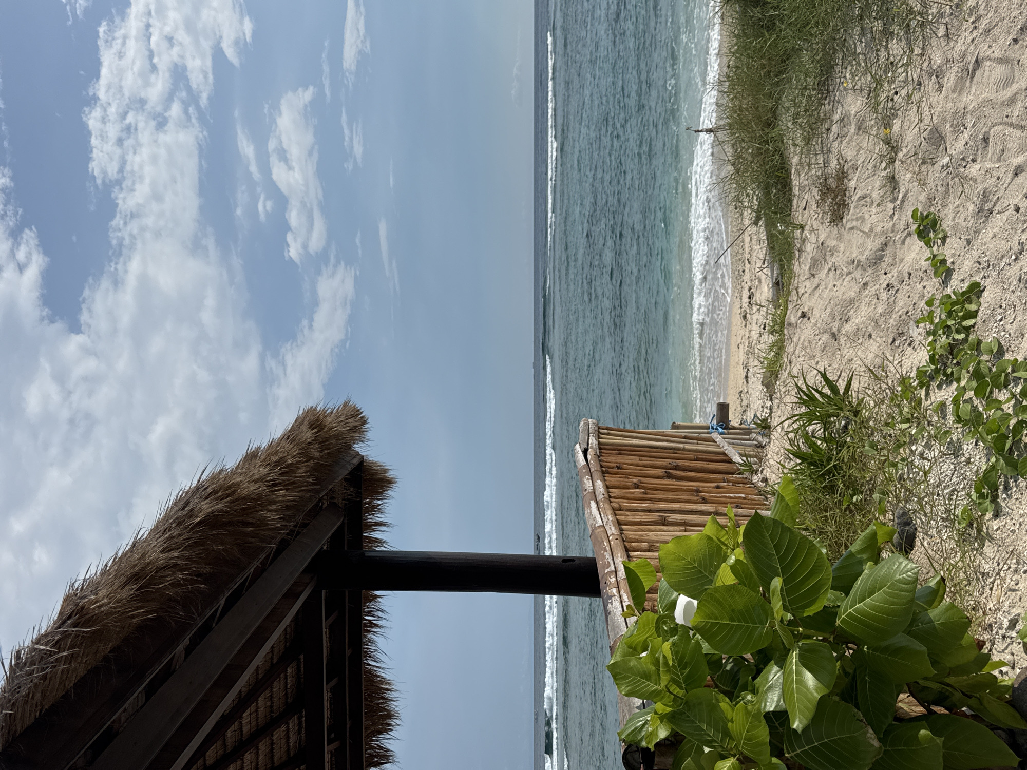 The scene is a beach in an unknown location in Indonesia. The image is bisected, showing the sky and sea. The left side features a partial view of a thatched roof structure. The right side presents a sandy beach with vegetation. Waves are visible, breaking on the shore. The sky is blue with scattered clouds.