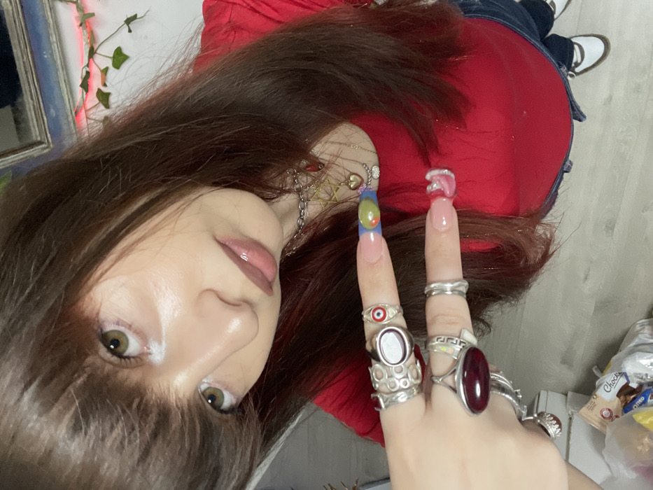 An image taken in an indoor setting within the city of Raanana, Israel, depicts a female subject from a high-angle perspective, likely a selfie. The subject has long, straight brown hair with bangs, fair skin, and greenish-brown eyes highlighted by white eyeliner on the lower lash line. She is wearing a red top, dark blue jeans, and white shoes with dark straps. Her left hand is prominently raised, displaying multiple silver rings, including an "evil eye" design on the index finger and a large oval red stone on the middle finger. Her nails are elongated and manicured; the left index finger nail features a blue base with yellow, green, and orange abstract patterns, while the left middle finger nail is light pink with a white swirl. Multiple necklaces are visible around her neck, including a silver chain with a Star of David pendant and another with a small heart pendant. The background consists of a light-colored floor and a white wall, with a partial view of a blue-framed object and artificial ivy vines adorned with small red lights. A white plastic bag containing packaged snack items is visible on the floor in the lower left. The subject is looking directly at the camera.