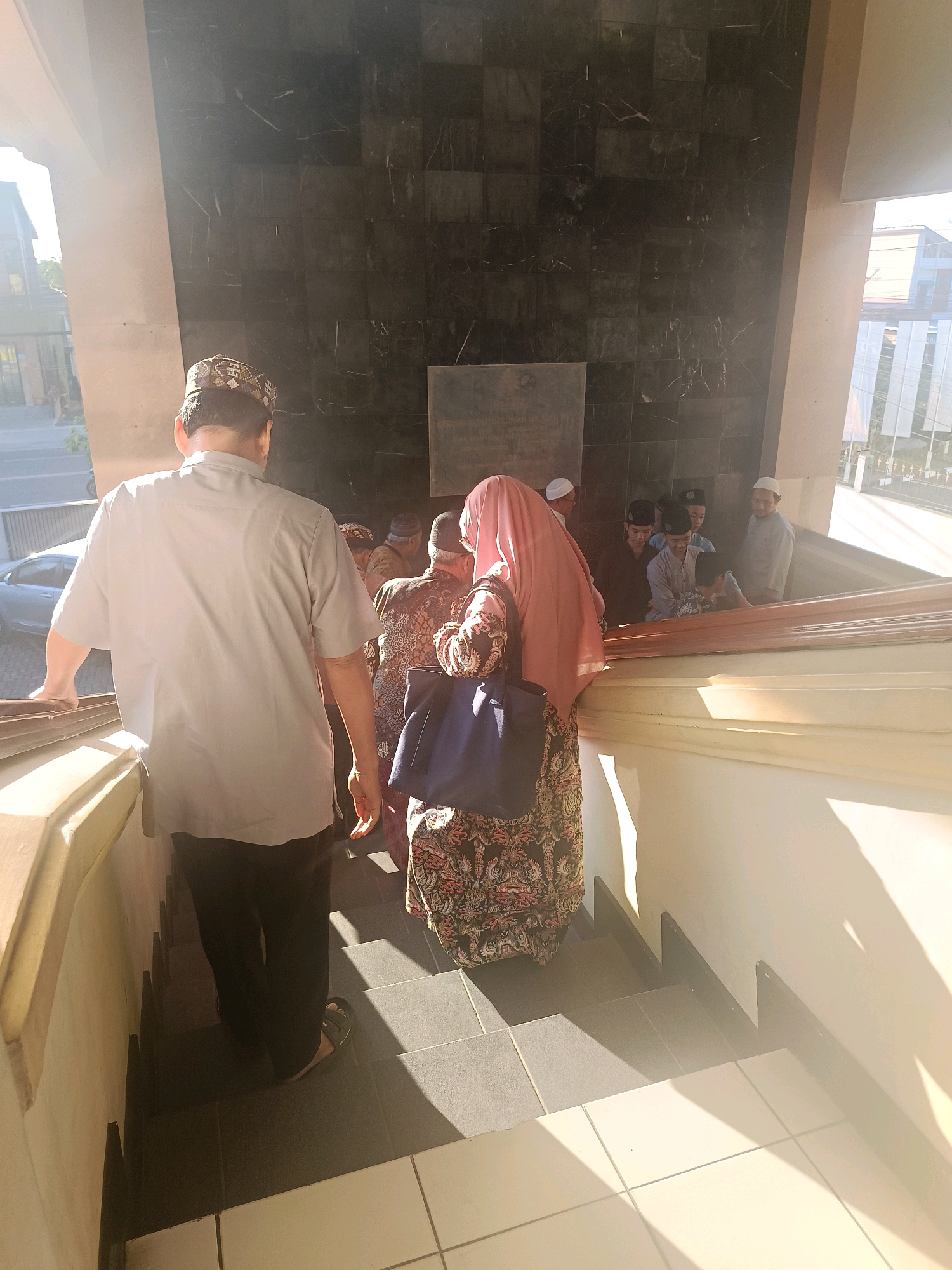 In Yogyakarta, Indonesia, a group of people, predominantly male, are descending a staircase. A man in the foreground, seen from behind, wears a light-colored short-sleeved shirt and dark trousers, along with a patterned skullcap. He is holding onto a railing. Behind him, a woman in a pink hijab and a patterned dress carries a dark blue bag. Several other individuals, also wearing skullcaps and traditional Islamic attire, are further down the stairs, some appearing to be younger. The staircase is made of dark gray tiles with lighter tiled risers, and the walls are covered in dark marble tiles. Sunlight streams in from the left, casting strong shadows on the stairs and walls. The background shows an exterior view with buildings and foliage.