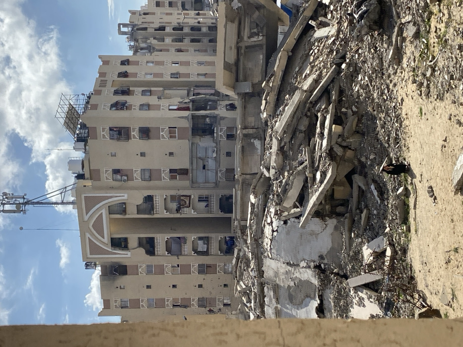 This image depicts a scene of widespread destruction in Khan Yunis, Palestinian Territory. In the midground, a substantial expanse of concrete rubble, rebar, and pulverized building materials from collapsed structures covers the landscape. Amidst this debris, a single individual, dressed in dark clothing and possibly wearing a head covering, walks on a sandy, cleared path. This person appears to be carrying a long, light-colored object diagonally across their left shoulder.
In the background, a large, multi-story beige building with distinct architectural elements, including a prominent arched decorative facade at the top center, stands partially damaged. The building exhibits visible signs of impact, including dark scorch marks, particularly on the left side of the upper floors, broken windows, and exposed interiors on multiple levels. Some balconies show signs of daily life, such as hanging laundry, while others are damaged. A cellular antenna tower is mounted on the roof of this structure. To the right and further back, additional damaged buildings are visible under a partly cloudy sky.