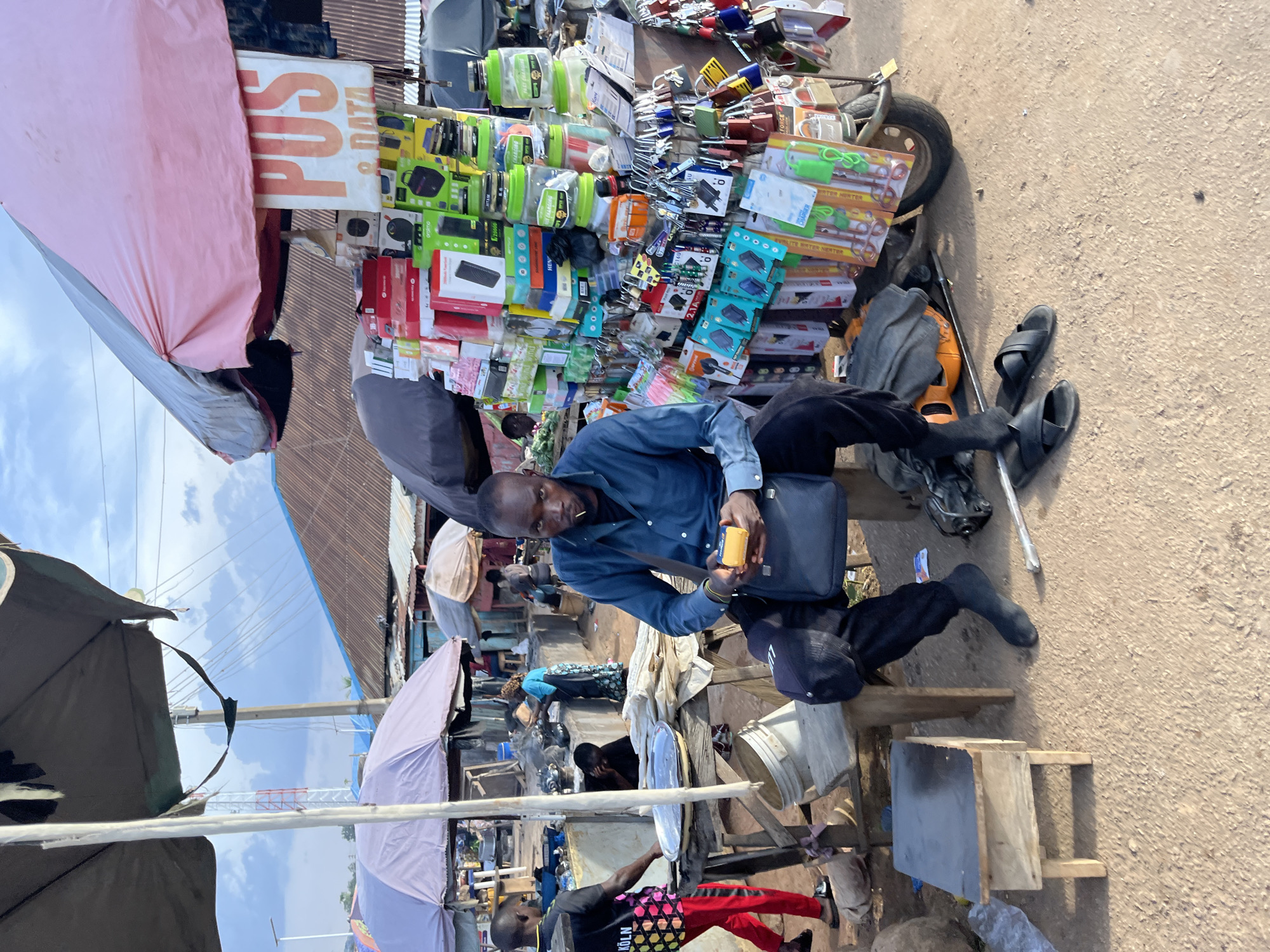 In Jos, Nigeria, a man sits by a stall displaying various electronics, including power banks, chargers, and phone accessories, all neatly arranged on a rack mounted to a tricycle. He wears a blue long-sleeved shirt and black pants, holding a small yellow device in his hands. A dark shoulder bag rests on his lap. The stall is covered by a pink and gray umbrella, and a sign above reads "POS & DATA". The background reveals a bustling market scene with other stalls, people moving about, and corrugated iron roofs of nearby structures under a cloudy sky. The ground is dirt and sand.