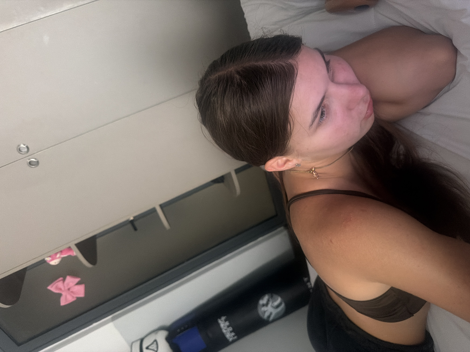 A young woman with long, dark hair lies on a bed, her head resting on her arm. She is wearing a dark brown bra and dark bottoms. Her eyes are downcast. Behind her, a white closet with silver handles is visible. To the left of the closet, a dark punching bag with blue and white accents hangs. On a shelf inside the closet, a pink bow and a small pink figure can be seen. The environment appears to be a bedroom.