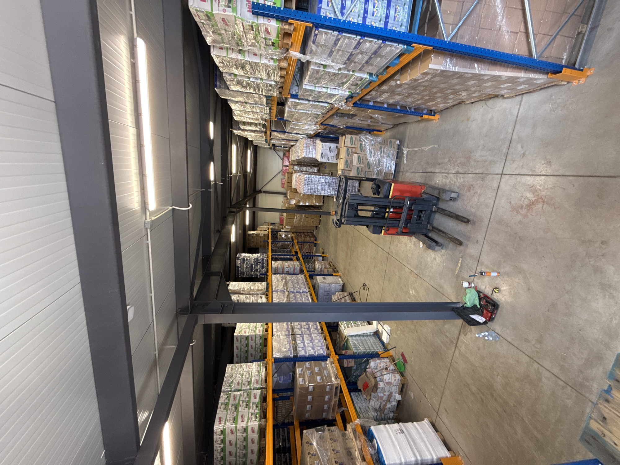 This is a medium-wide, eye-level, indoor shot of a warehouse in Graf Ignatievo, Bulgaria. The warehouse is filled with shelves stacked high with boxes of goods, wrapped in plastic. On the left side of the image, there are several tall shelves filled with boxes, with a large white stack of items at the bottom. In the center, a red forklift is parked, with a toolbox, several water bottles, and other small items scattered on the floor nearby. The floor is concrete, with visible seams. The ceiling is made of corrugated metal panels with fluorescent lighting. The overall impression is one of a busy, functional storage facility.
