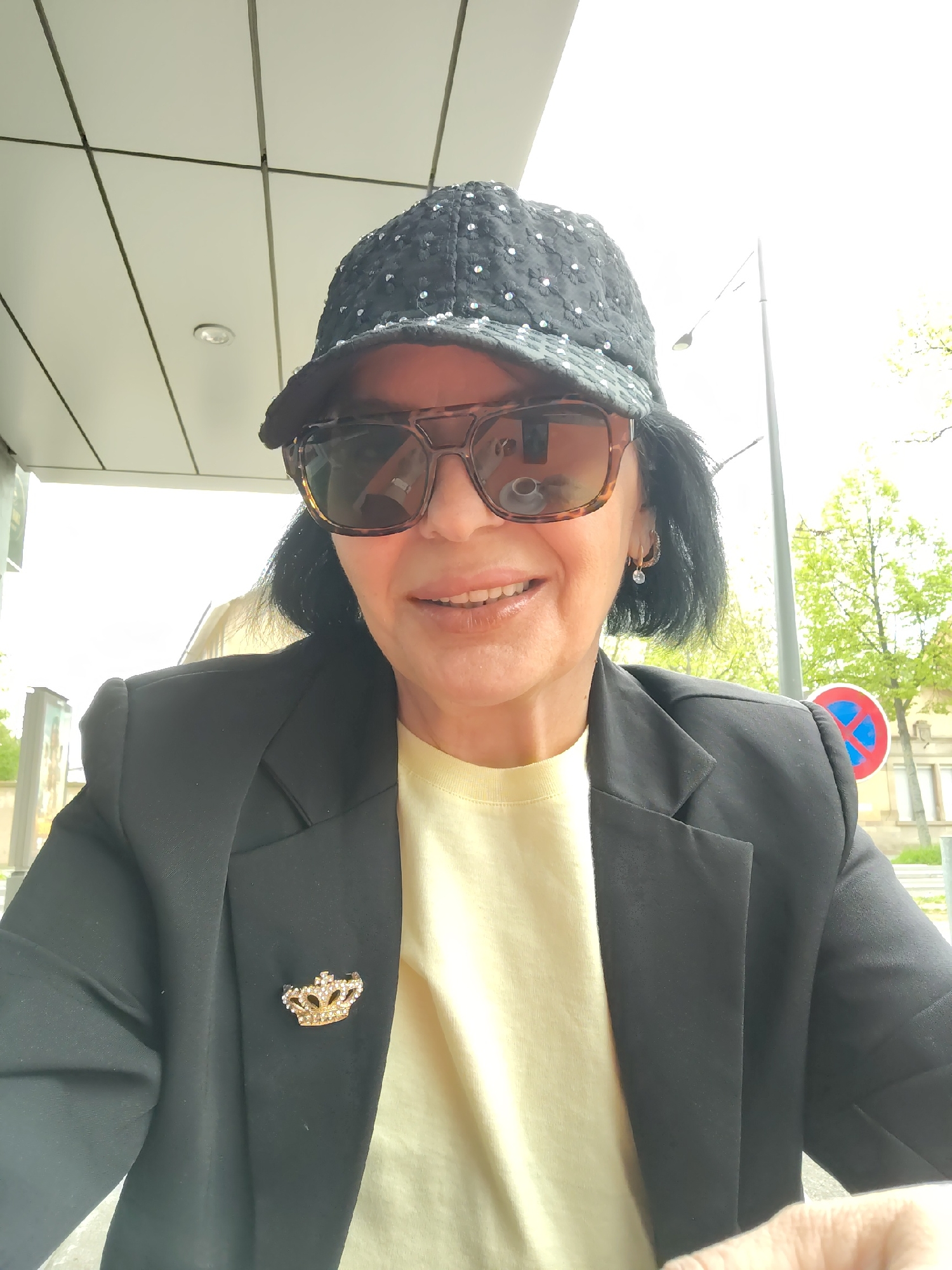 An older woman with dark hair wearing a sequined baseball cap and tortoise-shell sunglasses is posing for a selfie while sitting outdoors. She is wearing a black blazer over a light yellow shirt, and a small crown-shaped brooch is pinned to the blazer's lapel. A building with a white panelled exterior is visible behind her. In the background, across the street, are trees with green leaves and a blue and red traffic sign, indicating the location is likely Strasbourg, France. The scene appears to be a casual outdoor setting, possibly a cafe or restaurant.