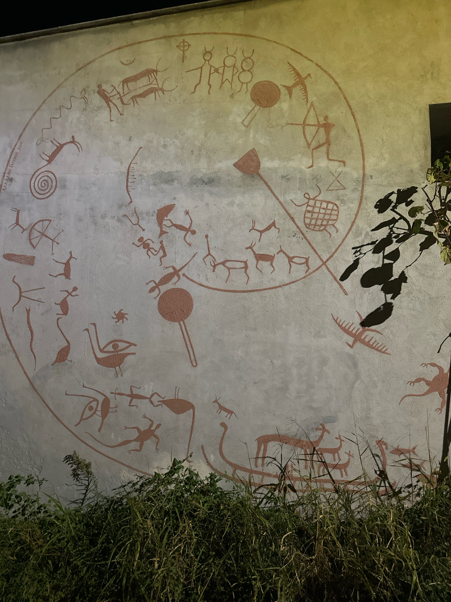 The image depicts a mural on a textured, light-colored wall, likely part of a building. The mural is rendered in a reddish-brown color and features stylized, ancient-looking pictographs of humans, animals, and abstract symbols arranged in a circular composition. The figures are simple, line-based drawings with minimal detail. Among the identifiable subjects are several deer-like animals, a bird in flight, humans in various poses including one with a bow and arrow, and what appear to be reptiles or amphibians. Some abstract shapes, including a spiral and a lattice-like pattern, are also visible. The overall style evokes prehistoric cave paintings. The mural is illuminated by an external light source, casting shadows from nearby foliage onto the wall. In the foreground, dense green vegetation and tall grasses obscure the lower portion of the wall. The upper right corner of the image shows a dark rectangular opening, possibly a window or doorway, and a hint of the building's structure. Text, appearing to be a name and date, is subtly visible on the left side of the mural, reading "АЛЕКСАНДАР ГАРУНОВ 2014". The mural is located on the wall of the "Mogren" fortress in Budva, Montenegro.