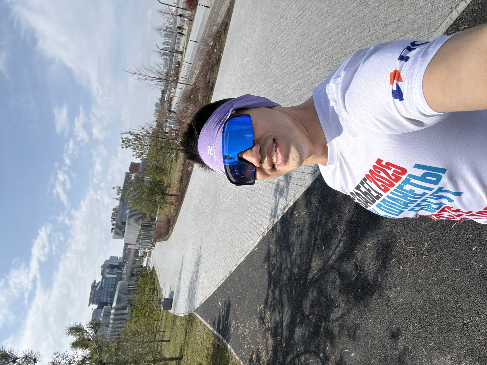 A man is taking a selfie outside on a sunny day in Kazan, Russia. He is wearing a white t-shirt with text in Russian and the year "2025" printed on it, a purple headband, and blue mirrored sunglasses. He is standing on a paved path that runs alongside a grassy area with small trees. In the background, there are modern buildings and a partially cloudy blue sky.