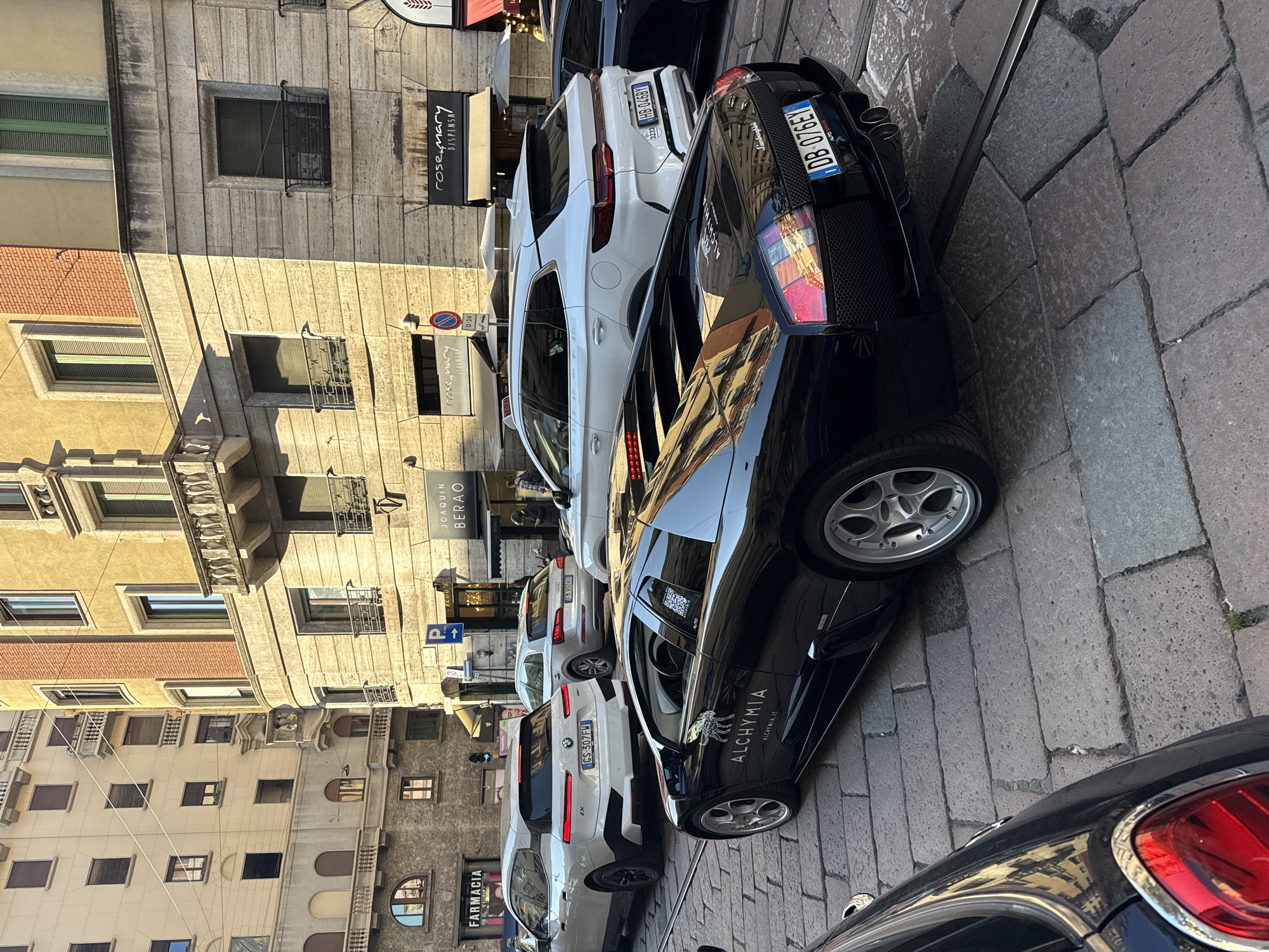 The image captures a vibrant street scene in Milan, Italy, featuring a mix of modern vehicles parked alongside historic, multi-story buildings under bright sunlight. The street itself is paved with cobblestones and includes tram tracks, indicating a busy urban environment.
Prominently featured is a black sports car, a Lamborghini, parked on the right side of the street, displaying the Italian license plate "DB-076EY". Its side bears the text "ALCHYMIA" and "ALCHYMIA.IT". Further down, a white BMW iX with license plate "GS-307FW" and a white Audi Q5 with "HB-046BY" are also visible. A dark vehicle with a distinctive red taillight is partially seen in the immediate foreground.
The surrounding buildings exhibit classic European architecture, characterized by stone facades, ornate balconies, and windows fitted with shutters or decorative grates. Visible storefronts and signs include "rosemary DISPENSA", "JOAQUIN BERAO", and "FARMACIA". A blue parking sign marked with a white "P" and an arrow, along with a "0-24" sign, suggests regulated parking.
The clear sky and strong, elongated shadows cast by the buildings and vehicles indicate that the photograph was taken during either the early morning or late afternoon. The overall atmosphere is that of a bustling, sun-drenched city street.