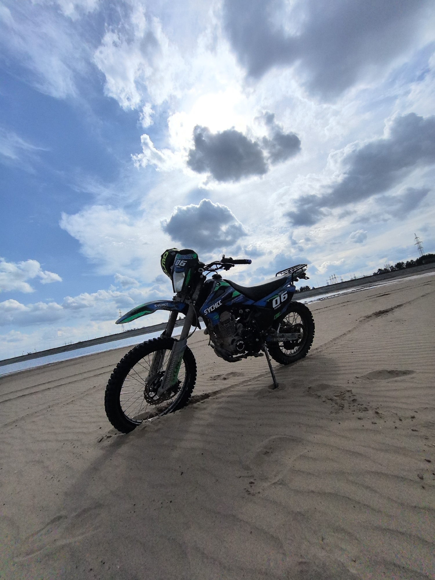 A blue and green dirt bike, identified by the brand "SYCHEE" and the prominent number "06" on its side panels and front, is parked on a sandy beach or shoreline. A black helmet rests on the bike's handlebars. The motorcycle features knobby tires suitable for off-road terrain, and its kickstand is extended into the sand.
The setting is an expansive sandy area, showing ripples and tire tracks, extending towards a body of water (likely a river or lake) in the mid-ground to the left. In the distant background, across the water, power lines and utility towers are visible, alongside a line of trees under a bright, partly cloudy sky. The sun, partially obscured by clouds, creates a strong glare in the upper right corner, indicating daytime, possibly late morning or early afternoon. The weather appears fair with a mix of clear blue sky and various cloud formations, from wispy to more substantial cumulus clouds.
The image captures a moment of rest during an outdoor excursion, likely off-roading, in a scenic, open landscape. No people are visible. The location context provided indicates this scene is near Chaikovsky, Russia.