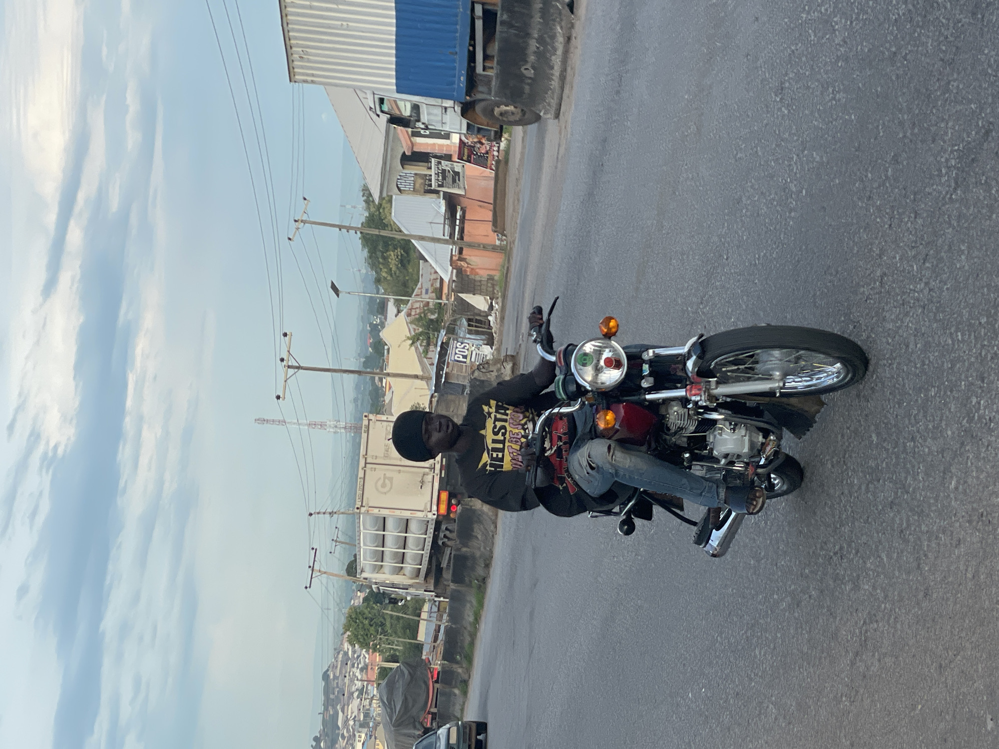 A young Black man wearing a black durag and a black t-shirt with yellow lettering "HELLSTAR MUST BE STORY" rides a red and black motorcycle on an asphalt road in Lokoja, Nigeria. He is wearing ripped blue jeans and dark sandals. The motorcycle has a round headlight and chrome accents. In the background, there are various buildings, trucks, and utility poles. One truck in the distance is white with a blue stripe, and another is a white tanker truck with a grey tarp. The sky is overcast with clouds. The scene appears to be on a highway or a busy road on the outskirts of the city.
