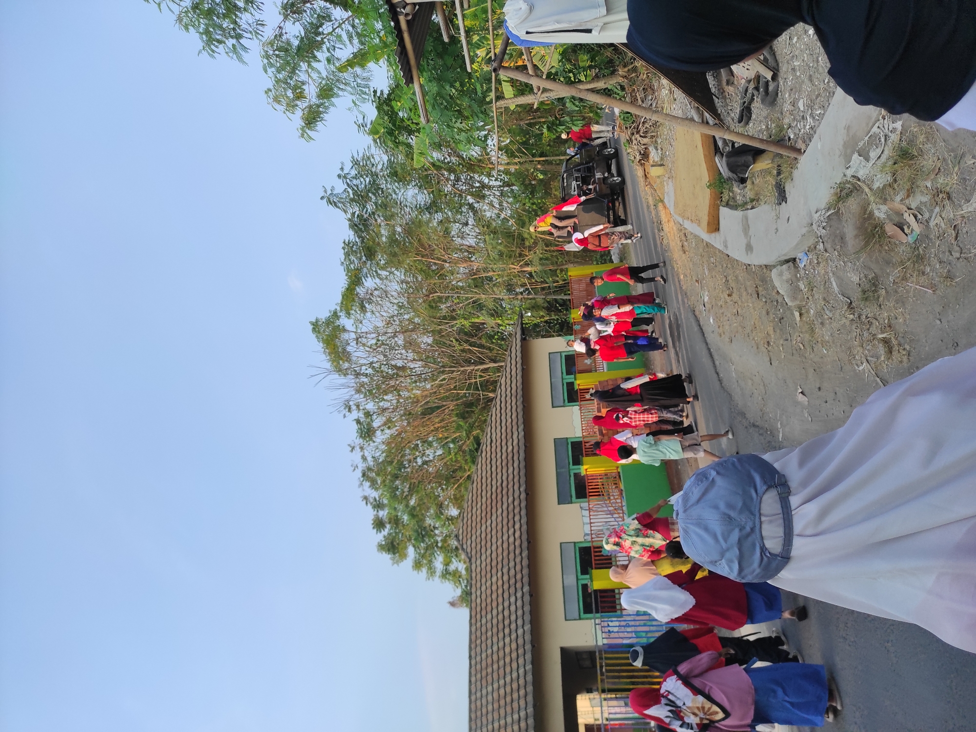 In Pleret, Indonesia, a group of people, some wearing hijabs and others in red and white attire, are walking along a paved road. The group appears to be part of a procession or celebration, with some individuals carrying flags. A black pickup truck is present, with people standing on and in it, possibly participating in the event. To the left of the road is a building with a tiled roof and colorful fencing, suggesting it might be a school or community center. Lush green trees and vegetation line the right side of the road, indicating a semi-rural or suburban setting. The sky is a clear, pale blue, and the lighting suggests it is late afternoon or early evening. The ground on the right side of the road is uneven and shows signs of construction or debris, including discarded materials and a wooden structure.