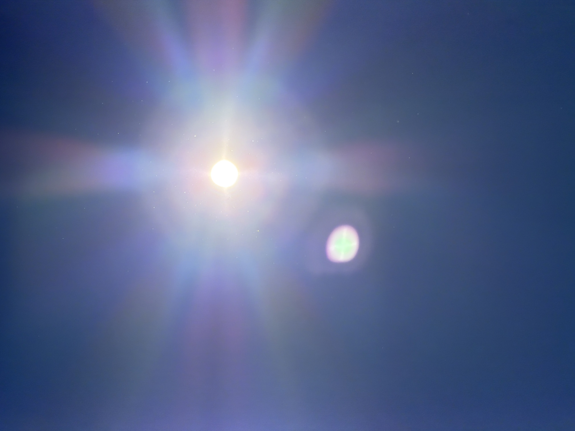 The image presents a bright light source, likely the sun, against a clear, deep blue sky. The sun, positioned slightly to the left of the frame's center, appears as an intensely bright white circular orb. Emanating from this central light are strong, multicolored light rays, creating a pronounced starburst or lens flare effect that stretches across a significant portion of the sky, displaying iridescent hues of purple, pink, and green.
To the right of the main light source, a distinct, out-of-focus, circular light anomaly is visible. This secondary light ghost is characterized by a blend of greenish and purple tones, indicative of further lens flare or internal reflections within the camera optics. Numerous tiny white specks are scattered across the entire blue sky, which could be attributed to dust on the camera lens or sensor, or perhaps very faint atmospheric particles.
The scene depicts a bright daytime setting with no clouds visible, suggesting clear weather. There are no human subjects, objects, or specific landmarks present to indicate a particular location within Bearpark, United Kingdom, aside from the provided context. The image captures the dynamic visual effects that occur when a camera is pointed directly at a powerful light source. No visible text is present in the image.