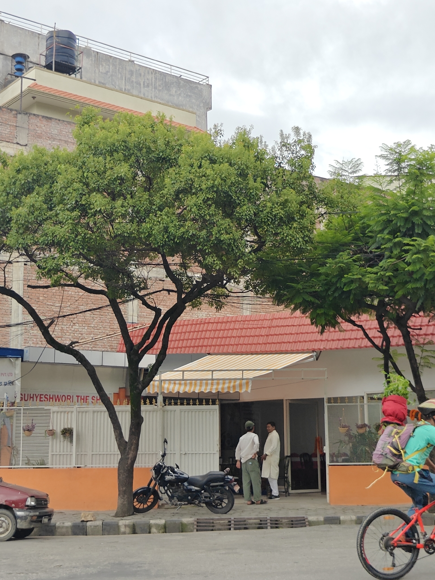 In Kathmandu Metropolitan City, Nepal, this image depicts a street scene with a building in the background. The building has a red tiled roof and an orange-painted lower section. There are several trees with green foliage in front of the building. A black motorcycle is parked on the sidewalk next to a tree. To the right, a person on a red bicycle wearing a helmet and carrying a backpack is riding past. Further to the left, a maroon car is partially visible. There are two men standing near the entrance of the building, one wearing a white shirt and trousers, and the other in a white kurta. A striped awning is extended over the entrance. The sky is overcast.
