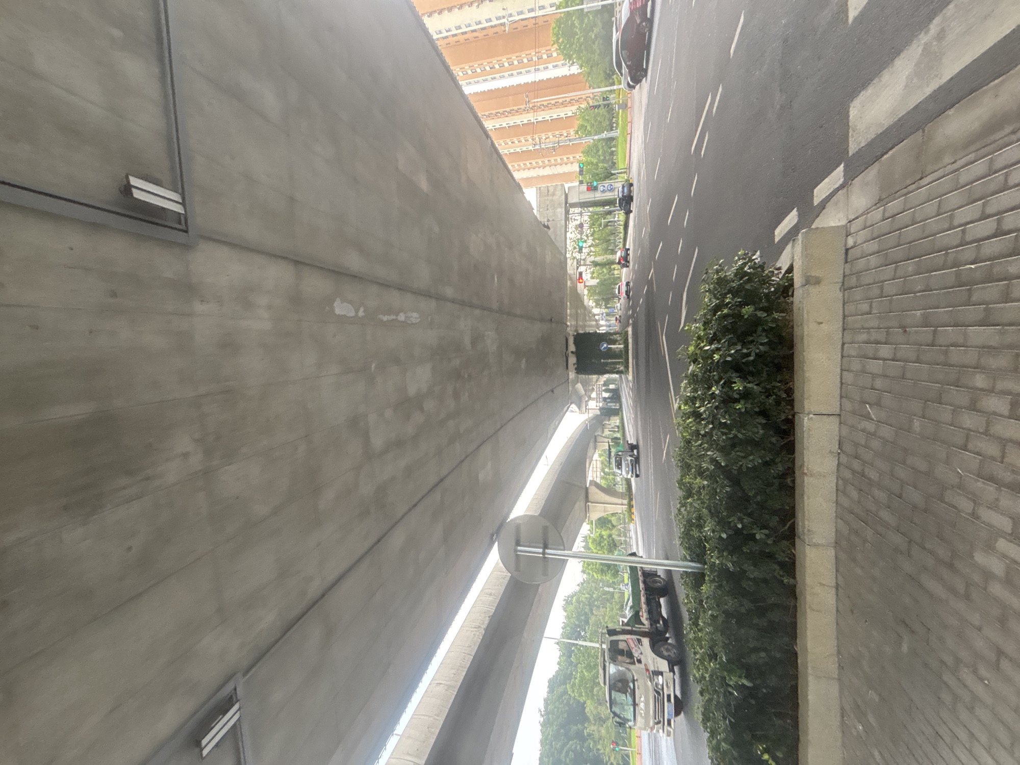 This is a wide-angle, low-angle shot of a concrete overpass in Suzhou Industrial Park, China. The overpass dominates the upper portion of the frame, with its textured concrete underside showing a repetitive pattern of support beams and embedded lighting fixtures. Below the overpass, a multi-lane asphalt road is visible, with clear white lane markings. Several vehicles are present: a white flatbed truck is in the left lane, a silver car is in the right lane, and other vehicles are further down the road. On the right side of the frame, a tall, orange residential building with numerous windows stands adjacent to the road. Lush green foliage lines the median and the edge of the sidewalk in the foreground, which is paved with grey interlocking bricks. Traffic lights are visible in the distance. The overall impression is one of urban infrastructure and daily city life.