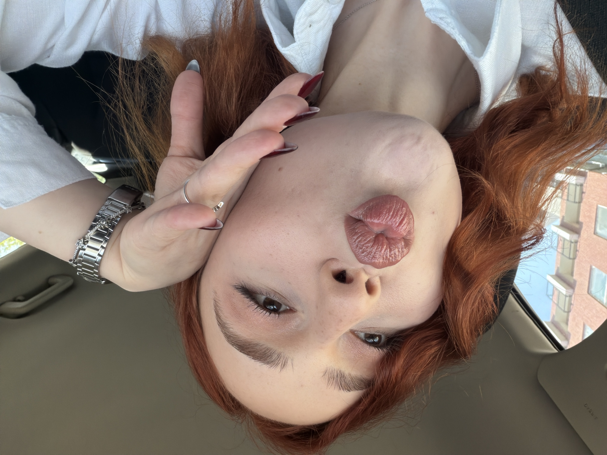 This image is an upside-down, close-up selfie featuring a person with fair skin, dark eyes, and long, wavy red hair. The individual's lips are pursed in a "kiss face" or pout, enhanced with glossy, muted pink lipstick. They are wearing a white collared shirt. Their left hand is positioned near their face, displaying manicured nails; some are dark red/burgundy, while at least one (thumb) is grey/blue-grey. A silver-toned watch with a link bracelet is visible on their left wrist, and a silver ring adorns their left ring finger.
The setting appears to be the interior of a car, with part of the light-colored car ceiling and a grab handle visible in the background. Through a window on the right side of the frame, a multi-story brick building and a clear blue sky are discernible, indicating it is daytime with sunny weather. The overall impression is a casual, posed selfie taken in an urban environment. No visible text is present in the media file.