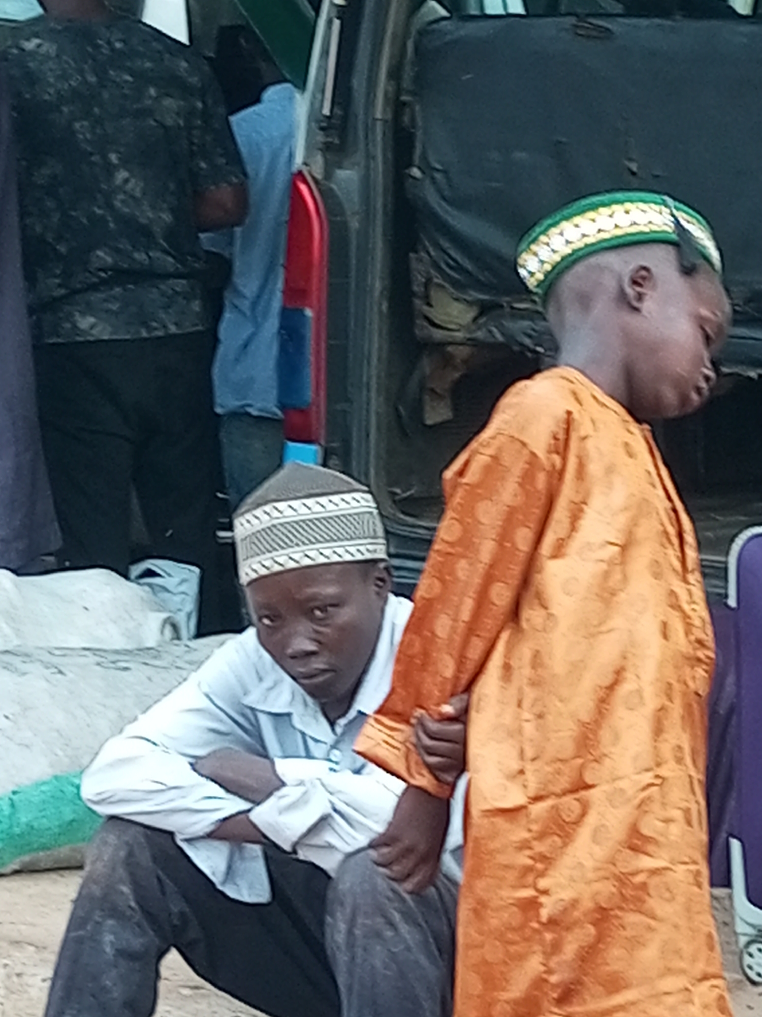 In Gombe, Nigeria, two young boys are seen near a vehicle. One boy, wearing an orange robe with circular patterns and a green and gold patterned cap, stands facing right with his head slightly down. The other boy, seated and wearing a white shirt and a patterned cap, faces the camera with his arms crossed and head tilted down. The background shows the interior of a vehicle, with black upholstery and a red and blue object visible. The ground appears to be dirt or sand, with some bags and a green cloth present.