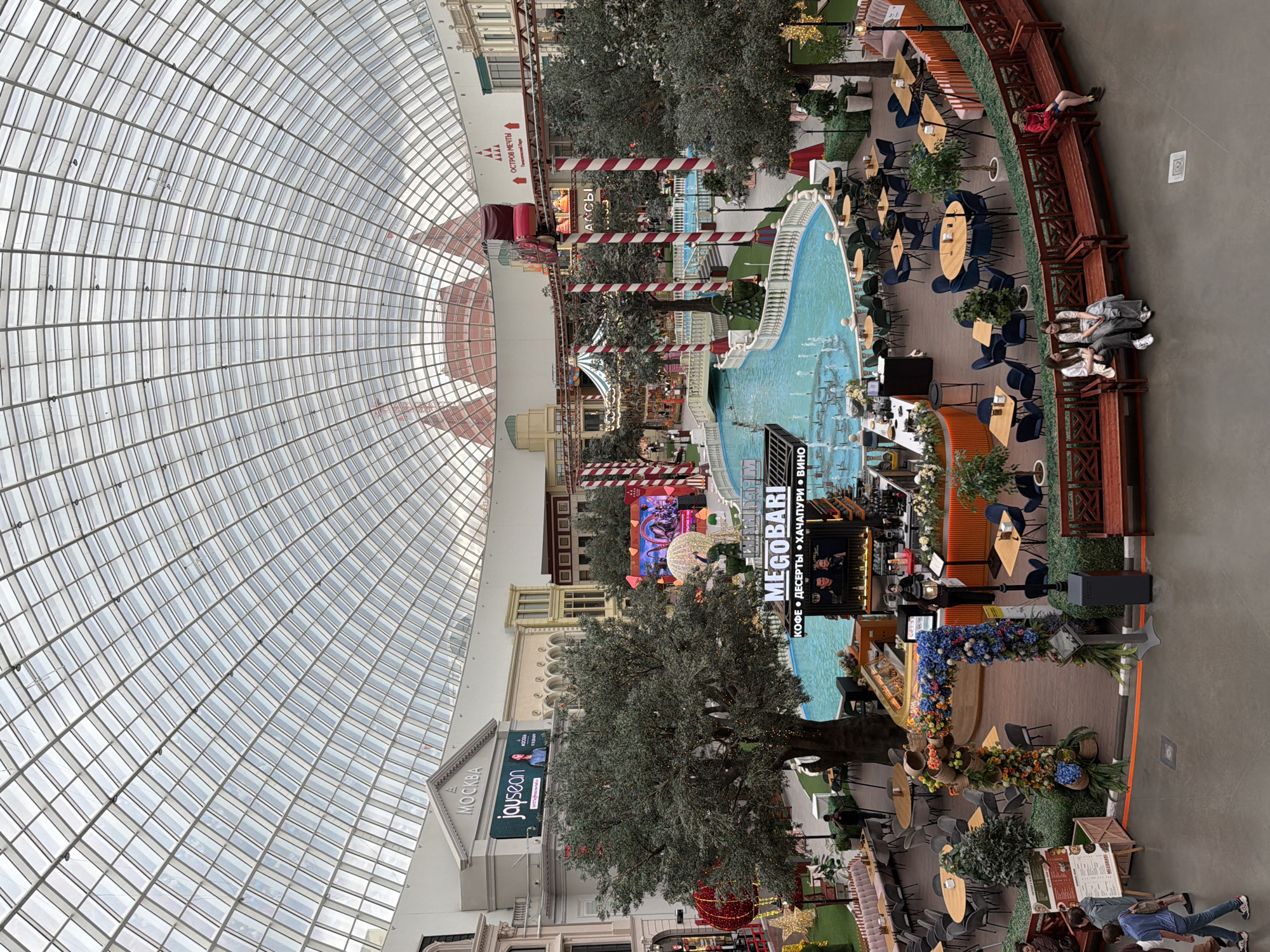 The image depicts an interior scene of a large building with a domed glass ceiling. The space appears to be a shopping mall or a leisure complex, featuring a central area with a blue swimming pool or decorative water feature surrounded by greenery, tables, and chairs, suggesting a café or restaurant setting. Artificial trees and floral decorations are strategically placed throughout the area, adding to the ambiance.
Several people are visible in the scene, some are sitting at tables, others are walking or sitting on benches. One person in a red jacket is reclining on a bench, while a group of three people are seated closely together on another bench. The overall atmosphere seems relaxed, with some individuals appearing to be socializing or taking a break.
A prominent sign reads "MEGOBARI" in large white letters, with smaller text below indicating "КОФЕ • ДЕСЕРТЫ • ХАЧАПУРИ • ВИНО" which translates to "COFFEE • DESSERTS • KHATCHAPURI • WINE," confirming the presence of a food and beverage establishment. Another sign in the background displays "МОСКВА" (Moscow). The lighting suggests it is daytime, possibly overcast, given the diffused light coming through the glass ceiling. The environment seems clean and well-maintained, with a mix of modern and decorative elements. The overall impression is one of a commercial and recreational indoor space designed for public access.