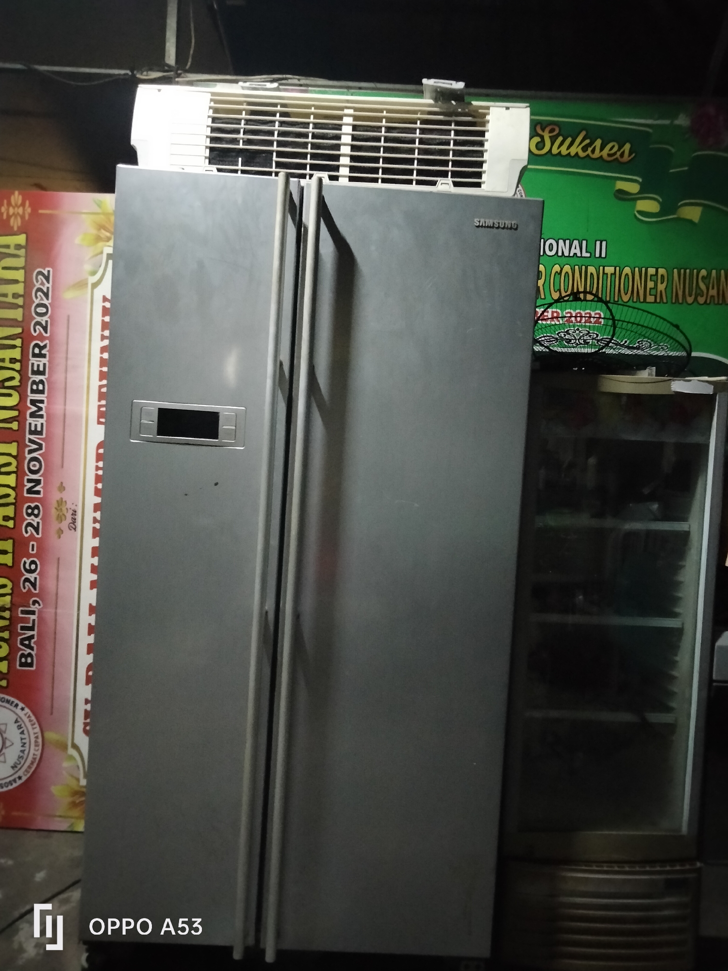 The image depicts a domestic scene, likely in Denpasar, Indonesia. Dominating the foreground is a large, silver, side-by-side refrigerator with a Samsung logo visible on its upper right door. The refrigerator appears to be in a somewhat cluttered environment, with an air conditioning unit positioned above it and to the left. To the left of the refrigerator, a portion of a red and yellow banner with text is visible, including the words "BALI, 26-28 NOVEMBER 2022". To the right of the refrigerator, a tall, glass-doored display cooler is partially visible. The lighting suggests it is either nighttime or an indoor setting with artificial lighting. The overall impression is one of a functional, everyday space, possibly a shop or a home storage area.
