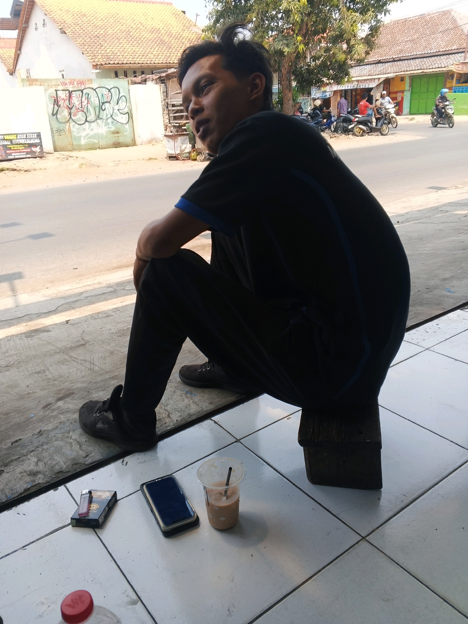 A person is sitting on a low wooden stool outside on a tiled floor. The person is wearing a black t-shirt with a blue collar and dark pants. Their legs are crossed. In front of them on the floor are a pack of cigarettes with a lighter on top, a smartphone, and a plastic cup with a straw containing a beverage. Behind the person, a street is visible with several motorcycles and buildings. The overall scene appears to be in a commercial or residential area.