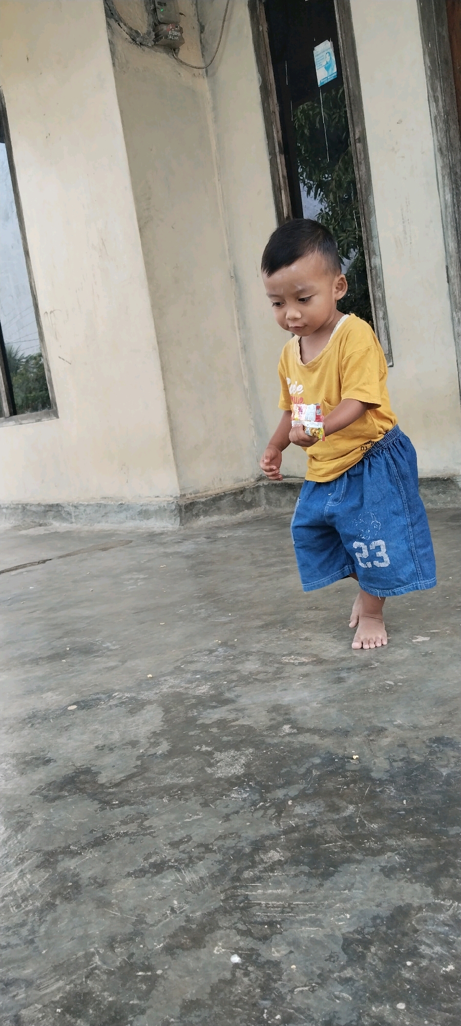 In Tanjung Ratu Ilir, Indonesia, a young boy stands on a concrete ground. He is wearing a yellow t-shirt and blue denim shorts. The boy is holding a small package in his right hand, appearing to examine it. His left hand is extended slightly downwards. Behind him, a building with a cream-colored wall and a window with wooden frames is visible. The ground is wet and shows signs of discoloration. The overall scene suggests an outdoor environment near a residential structure.