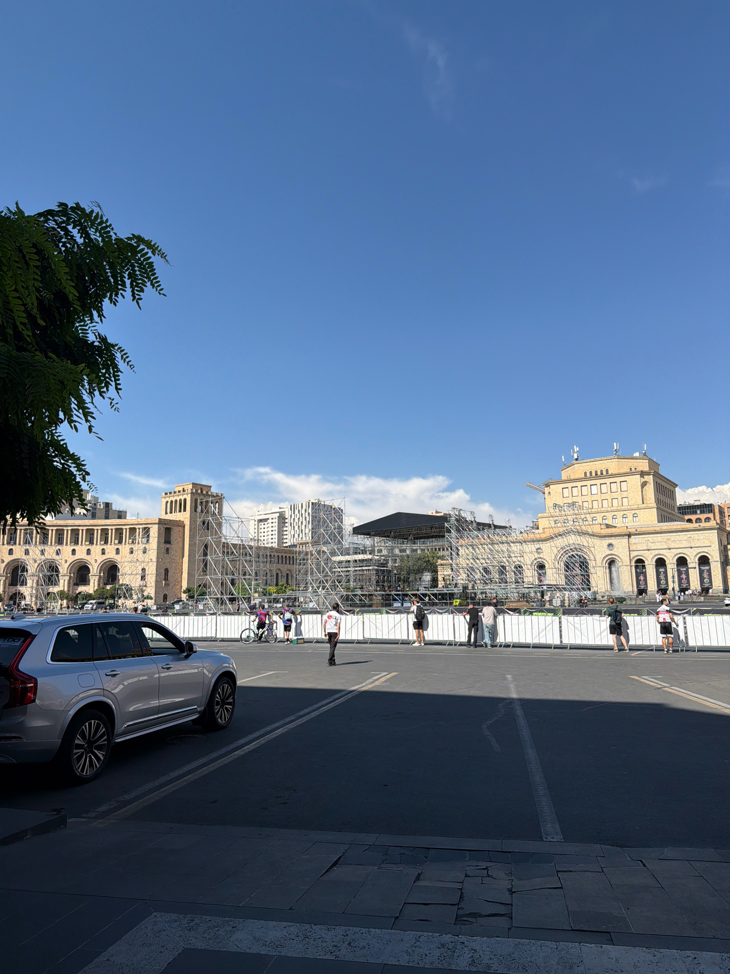 A silver Volvo XC90 is parked on the left side of the frame, partially in shadow. In the background, a large, open plaza is visible with a clear blue sky overhead. Several multi-story buildings with classical architecture are present, some featuring scaffolding. There are people walking and gathered in the plaza, and a white barricade separates the plaza from the street. The scene appears to be a public square in Yerevan, Armenia, possibly undergoing preparations for an event.