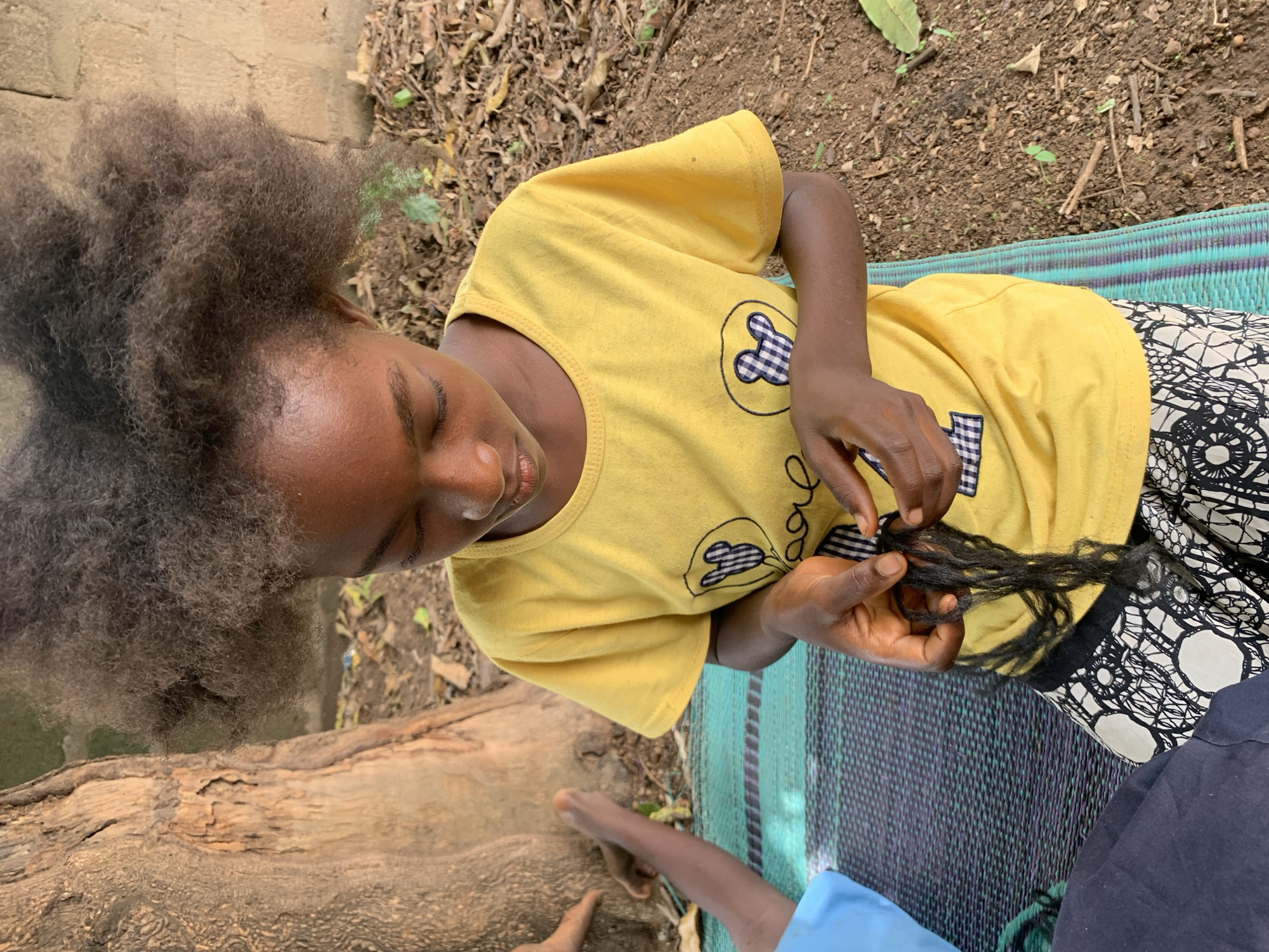 Akwanga, Nigeria. A brown-skinned person with dark, voluminous hair is seated on a patterned mat outdoors, wearing a yellow t-shirt with cartoon bear images. The person is holding and manipulating several strands of dark hair. The background includes foliage, a log, and bare earth.