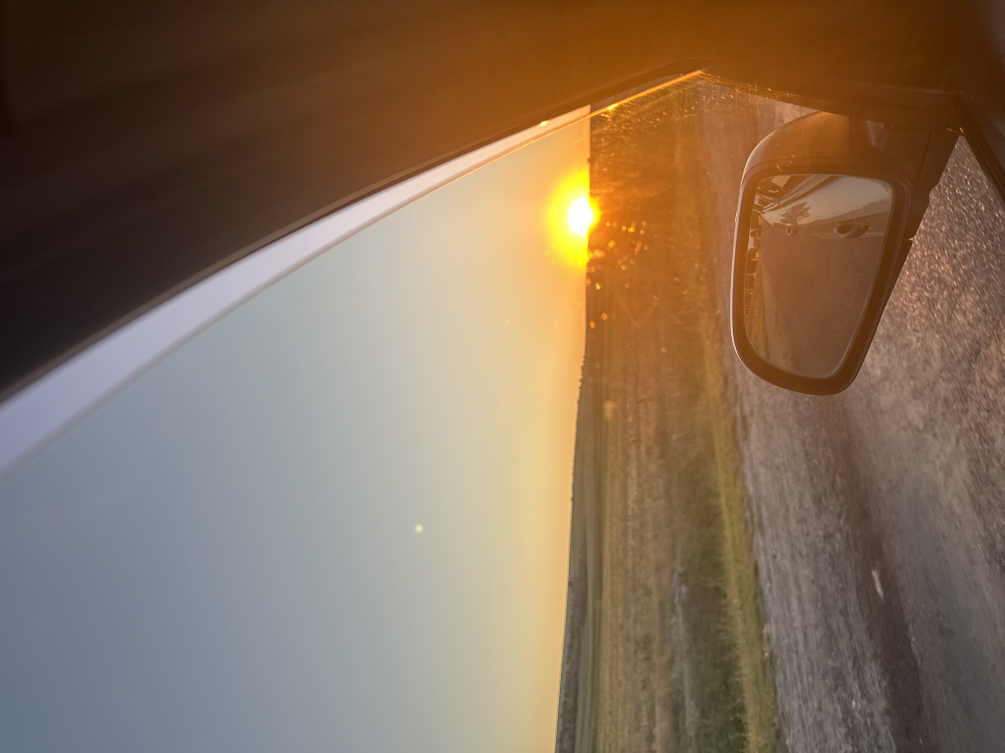 This image depicts a roadside view from inside a vehicle, capturing a sunset over a rural landscape. The sun, a brilliant orange sphere, is just beginning to dip below the horizon, casting a warm glow across the scene. Fields of dry grass stretch out to the left, bordered by a faint treeline in the distance. A dirt road runs parallel to the fields. A car's side mirror is visible in the foreground, reflecting the road and the car's interior. The sky transitions from a pale blue at the top to a vibrant orange near the horizon. The image appears to be taken in a rural area, possibly on a road leading out of or into Cioc-Maidan, Moldova.