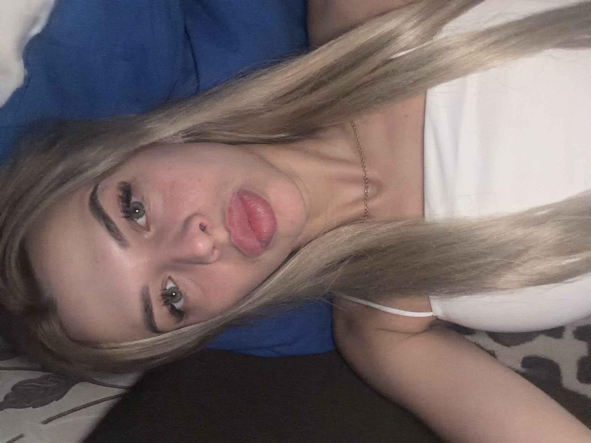 A young woman with blonde hair is lying down, looking at the camera. She is wearing a white top and a thin chain necklace. Her lips are slightly pursed. The background is a blurred mix of dark and blue fabric, possibly a bed or sofa. There is no discernible information about the location being Kyiv, Ukraine.