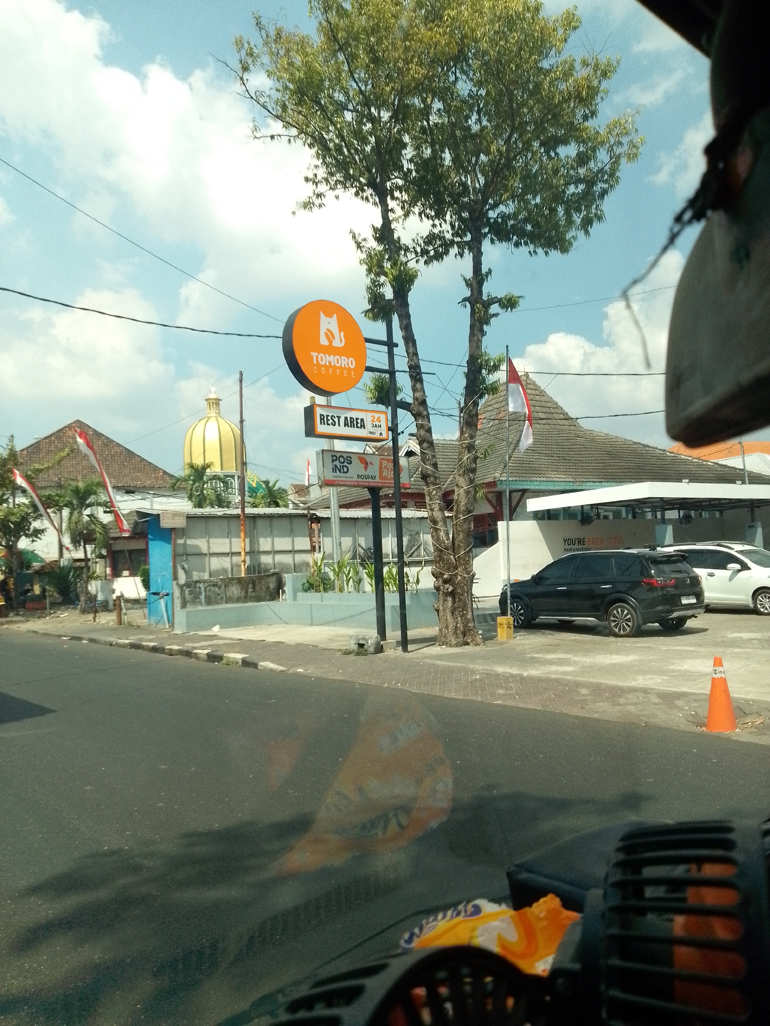 In the city of Babat, Indonesia, an outdoor scene unfolds under a bright, partially cloudy sky. A black SUV and a white car are parked in front of a building with a gray roof and white awnings. A sign with a circular orange logo and the text "TOMORO COFFEE" is visible, along with another sign indicating "REST AREA 24 JAM". An orange traffic cone stands on the side of the road. In the foreground, the interior of a vehicle is partially visible, with a dashboard and what appears to be a fan or vent. Shadows from trees and possibly the vehicle are cast on the asphalt road. The environment appears to be a roadside establishment, possibly a rest stop or coffee shop.