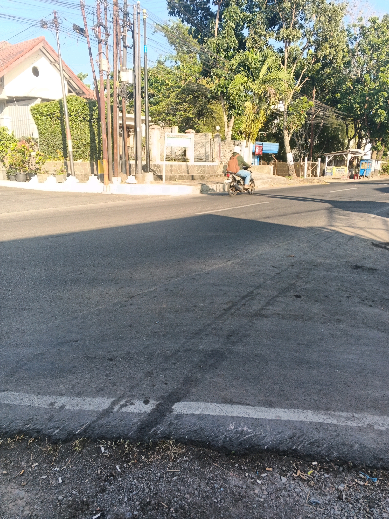 The image shows a residential street in Semarang, Indonesia, with a person riding a motorcycle. The street is paved with asphalt and has a white line marking the lane. On the left side of the street, there is a house with a red tile roof and a neatly trimmed green hedge. Utility poles with numerous wires are visible. On the right side of the street, there are trees, a small covered structure that appears to be a kiosk or food stall, and a blue trash bin. The sky is clear and blue, indicating a sunny day. The motorcycle is being ridden by a person wearing a helmet and a backpack.