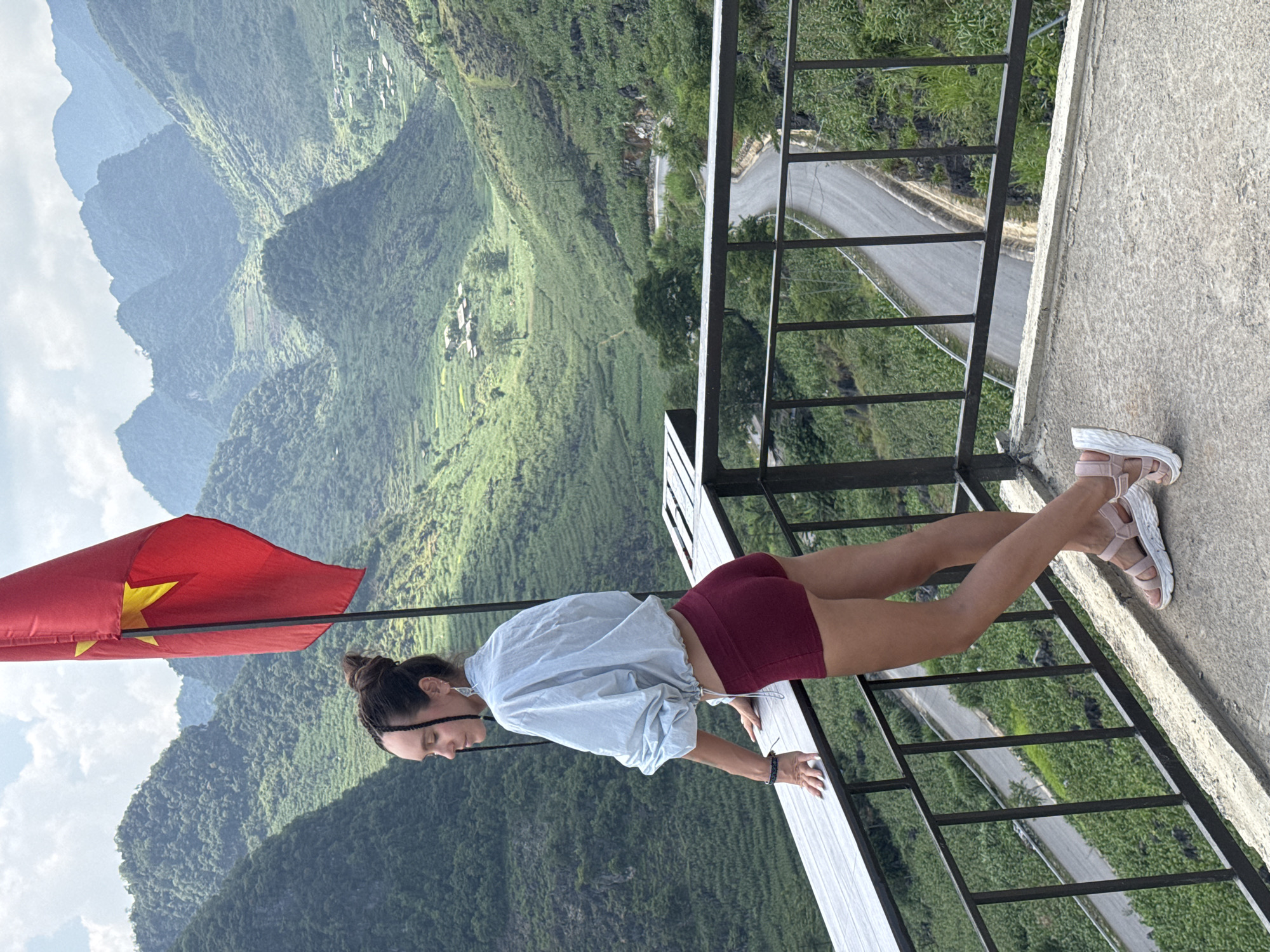 A woman stands on a balcony in Lũng Phìn Commune, Vietnam. She is wearing burgundy shorts, a light blue shirt, and pink sandals. She is holding onto the railing of the balcony. In the background, there are mountains, trees, and a winding road. The Vietnamese flag is visible in the distance.