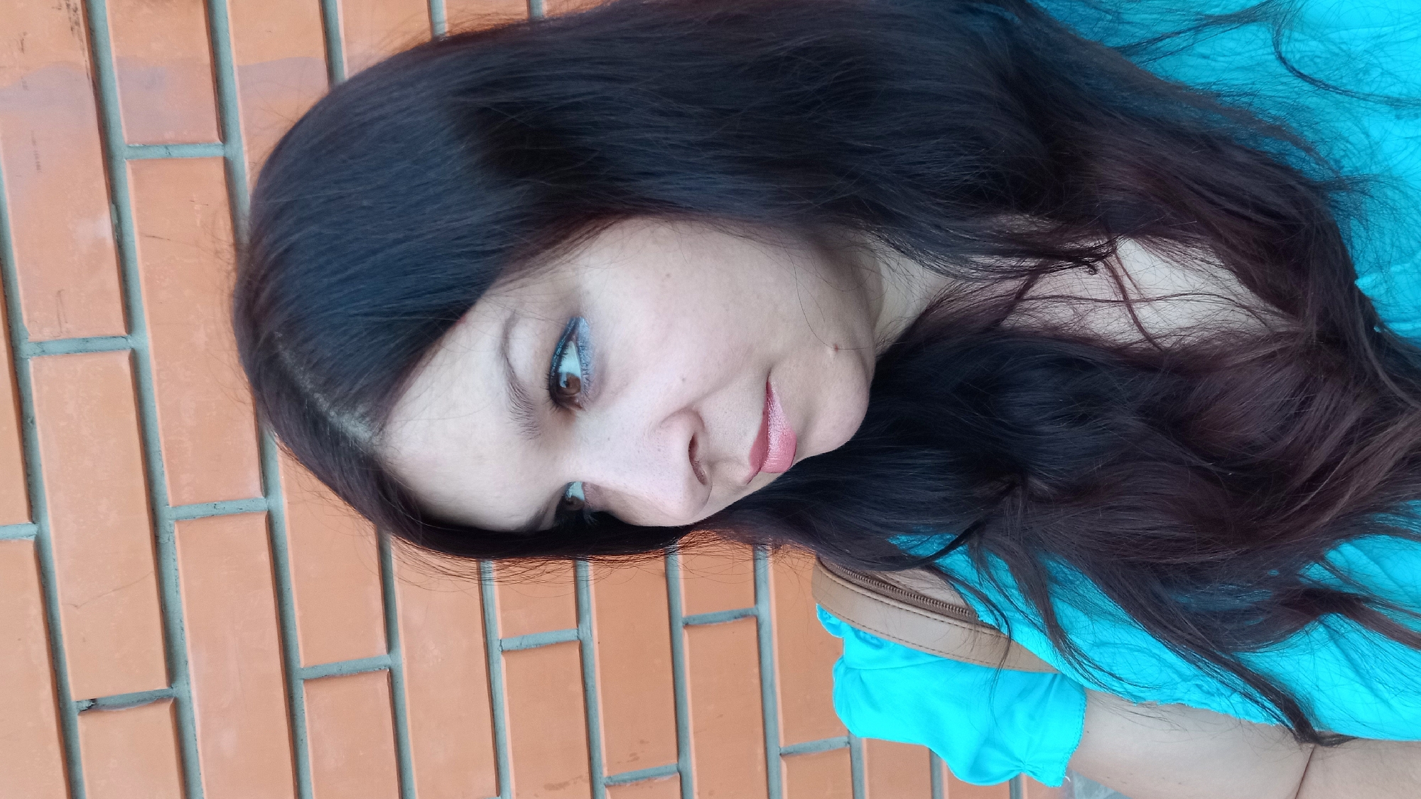 A woman with dark brown hair is looking to her left, captured in a medium shot against a brick wall. She is wearing a bright turquoise top and a brown strap of a bag is visible across her shoulder. Her makeup includes light eyeshadow and pink lipstick. The brick wall behind her is made of orange bricks with visible mortar lines. The scene appears to be outdoors, possibly on a street or in front of a building in the city of Kropyvnytskyi, Ukraine.