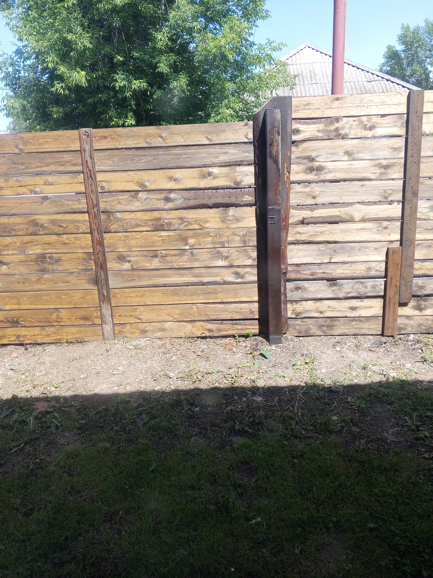The image shows a weathered wooden fence with horizontal planks. A dark, vertical metal post is affixed to the fence on the left side, and a narrower wooden plank with nails is attached to the fence on the right side. The fence is situated in an outdoor environment with a patch of dirt and sparse vegetation in front of it, and lush green grass further in the foreground. Behind the fence, a large green tree with dense foliage is visible, and beyond that, a house with a corrugated roof and a red pipe. The sky is clear and blue. The scene is likely located in the city of Maslyanino, Russia, given the architectural style and vegetation.