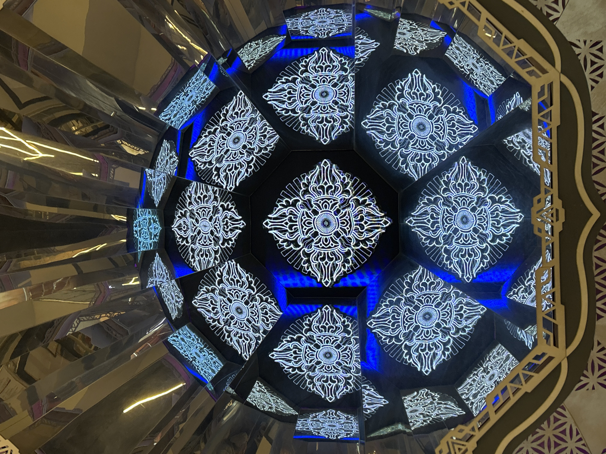 The photograph presents a close-up view of a decorative structure, possibly part of an interior or exterior design element. The structure consists of a rounded, segmented surface, where each segment is a polygon with a dark background featuring an ornate, symmetrical white pattern that is illuminated. Blue light appears to be emanating from the spaces between the polygonal segments. The structure has reflective, metallic elements visible on the left side of the image. The right side is partially obscured by a dark brown and beige ornamental design, possibly a frame or architectural detail. The city and location are unknown.