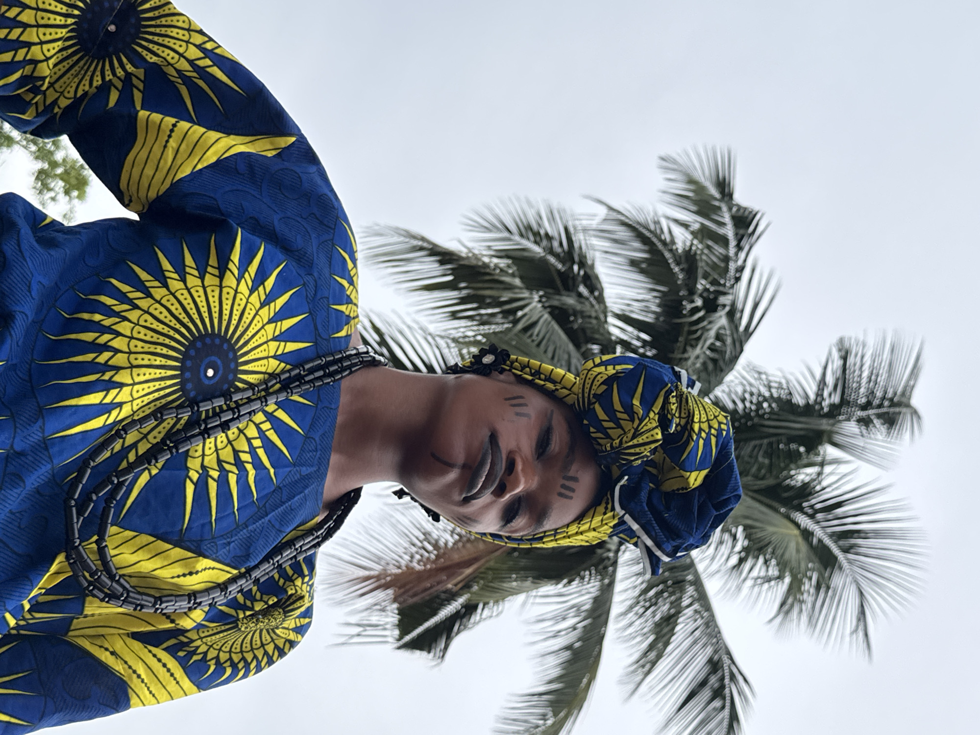 A low-angle medium shot shows a Black person in a blue and yellow African print outfit. They are wearing a matching headwrap. Black linear designs are painted on their face. They are wearing a multi-strand beaded necklace. A palm tree is in the background. The location is Badagry, Nigeria.