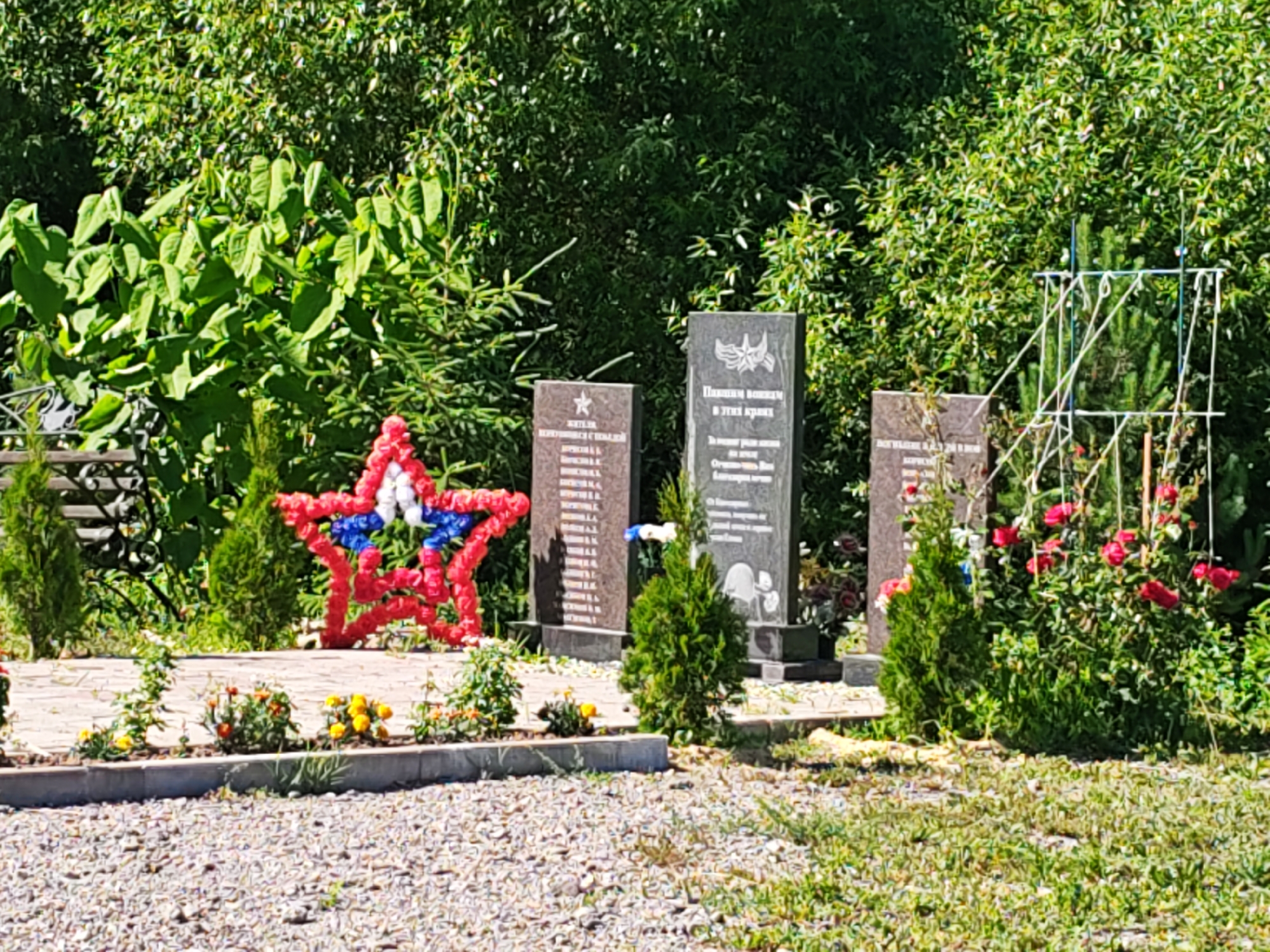 This is a ground-level, eye-level, slightly wide shot of a war memorial. In the foreground, a bed of gravel is visible, transitioning to a paved walkway lined with low-growing flowers in yellow and orange hues. To the left of the walkway, a red, white, and blue star-shaped floral arrangement is prominently displayed. Behind the flowers, a stone bench is partially visible.
The main focus of the image consists of several gravestones or monuments. The first monument on the left is a dark gray rectangular slab with an inscription and a star symbol at the top. To its right is a larger, taller monument of similar material, featuring an inscription and a depiction of wings. Further to the right, another monument of the same type is visible. In front of these monuments, several small, conical evergreen trees are planted.
The background is dominated by lush green foliage, appearing as dense bushes and trees, suggesting a park or garden setting. The sky is not visible in this frame. The overall impression is one of solemn remembrance and tribute.