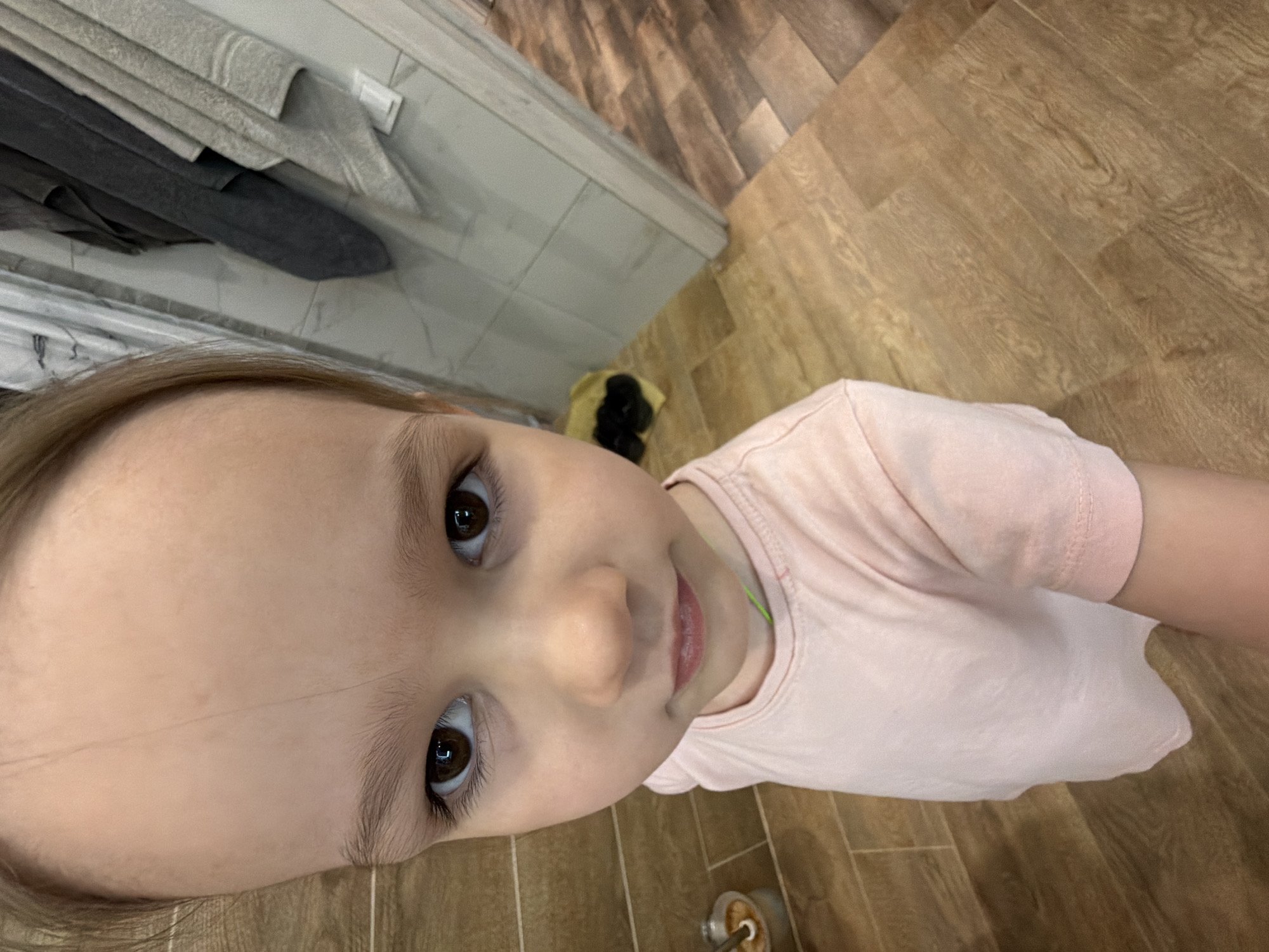 A young girl is looking up at the camera. She has brown eyes and is wearing a light pink shirt. The background shows a tiled floor and a wall, suggesting an indoor setting, likely a bathroom or hallway. It is difficult to ascertain the location as Chaikovsky, Russia without more contextual clues from the image.