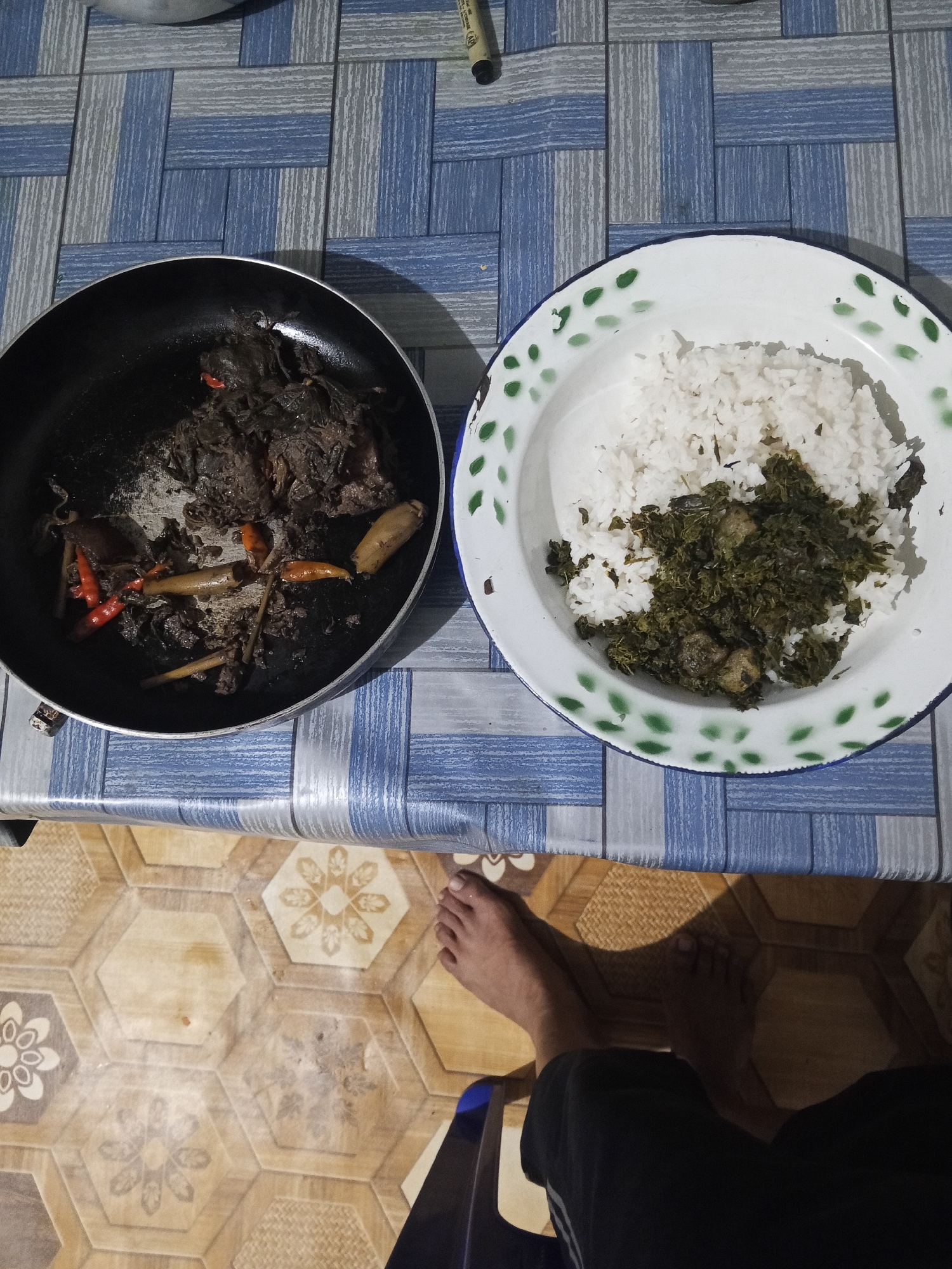 In Makale, Indonesia, a meal is served on a table covered with a blue and white checkered tablecloth. To the left, a black frying pan contains dark-colored food, possibly meat, with visible red chili peppers and ginger stalks. To the right, a white enamel plate with a green patterned rim holds a portion of white rice topped with a dark green leafy vegetable and chunks of meat. The floor beneath the table has a hexagonal pattern with floral and textured designs. A person's feet and legs, clad in black shorts, are visible in the lower right portion of the image, suggesting they are seated at the table. A shadow from a nearby object falls across the floor.