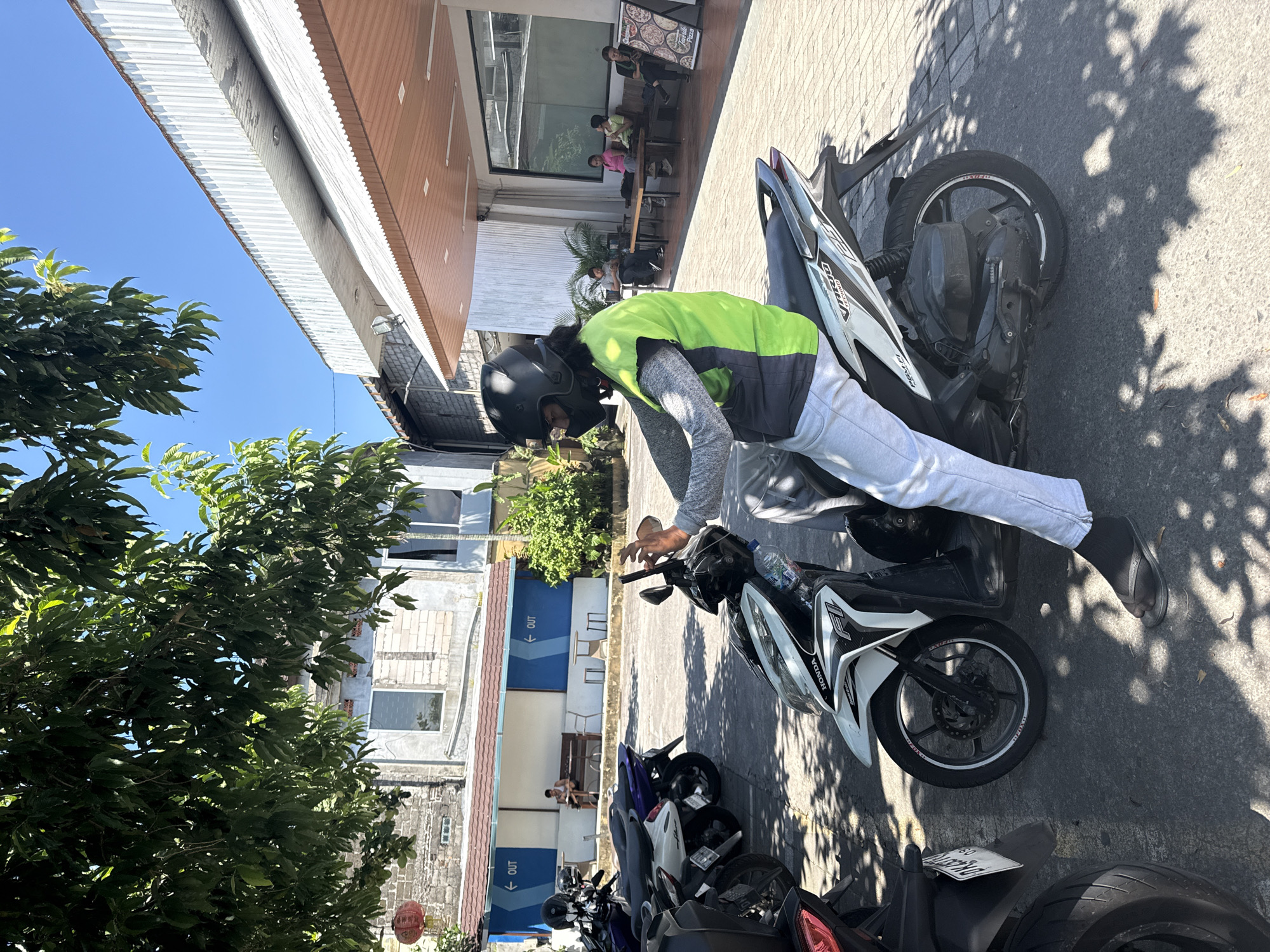 A man wearing a black helmet, light gray pants, and a green and gray long-sleeved shirt is straddling a white Honda motorcycle in the city of Petulu, Indonesia. Several other motorcycles are parked behind the one he is on. A building with blue "Out" directional signs is visible in the background, along with a tree and another building with people seated inside near a glass window. Shadows from the tree are cast on the ground.