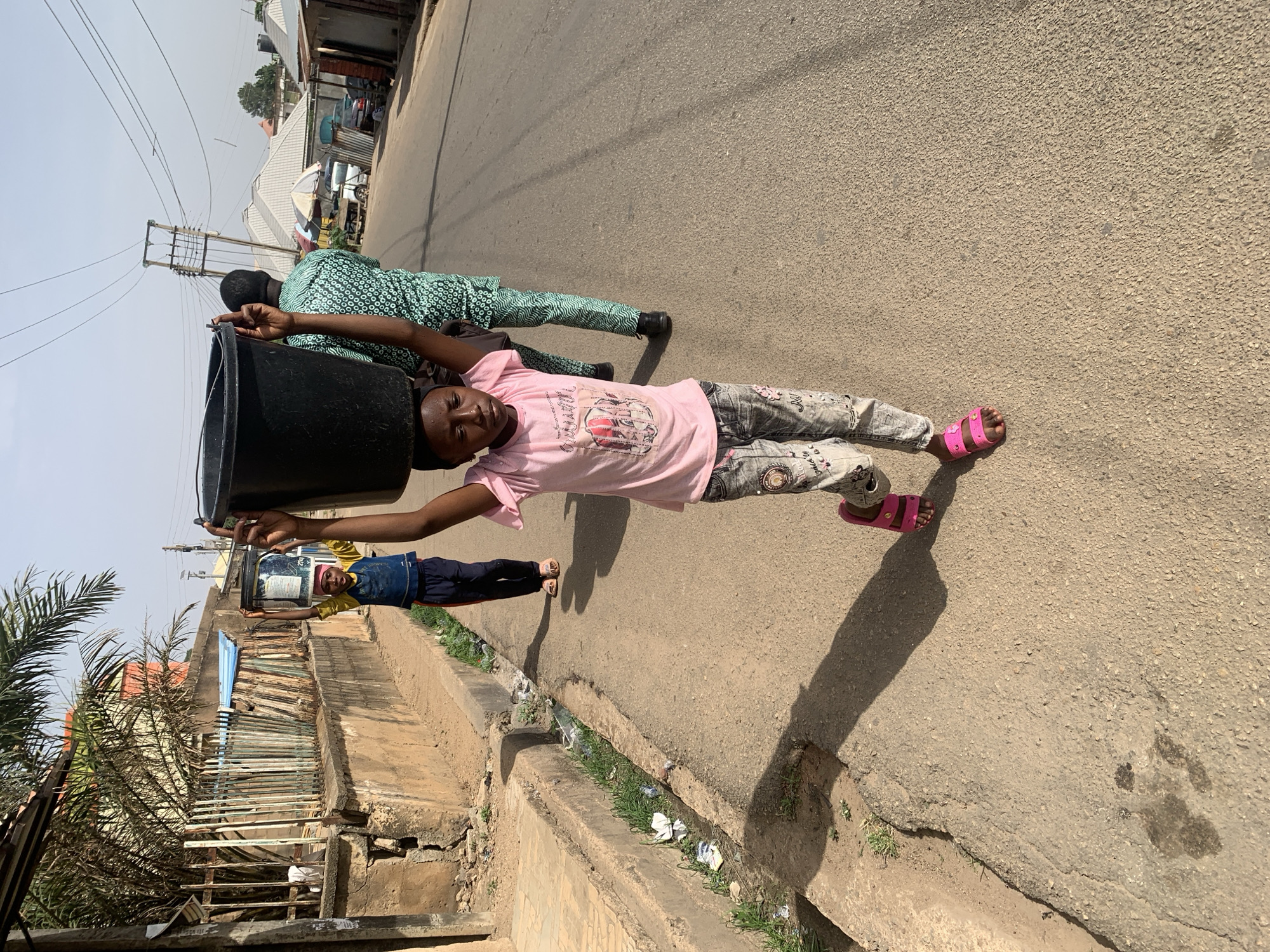 The image depicts a bright, sunny street scene in Jos, Nigeria, capturing three individuals walking along a paved road, all carrying buckets. The photograph is rotated 90 degrees clockwise.
In the foreground, a young girl, possibly a child or pre-teen, is shown looking directly at the camera with a neutral expression. She wears a pink short-sleeved t-shirt with a graphic print, grey patterned trousers, and bright pink open-toed sandals. She balances a large black bucket on her head, steadying it with her right hand.
Behind her, another individual is seen from the back, wearing a green long-sleeved top and matching trousers with a circular pattern. Further down the road and to the left, a third, smaller individual, likely another child, also walks while carrying a bucket on their head. This child is wearing a yellow top and blue trousers.
The setting is a paved asphalt road, showing some wear and cracks. Strong sunlight casts distinct shadows of the people onto the road, indicating it is likely mid-day or early afternoon with a clear sky. Along the left side of the road, a concrete drainage ditch contains some grass and scattered litter. Beyond the ditch, various structures resembling simple fences and buildings, possibly residential or small businesses, line the street. Utility poles with power lines crisscross the upper portion of the frame. Palm trees and other vegetation are visible near the buildings.
The activity clearly shows daily life, with the individuals engaged in transporting buckets, possibly for fetching water or carrying goods. No clearly legible text is visible on any clothing or objects, though there are graphics on the pink t-shirt and faint markings on one of the buckets.