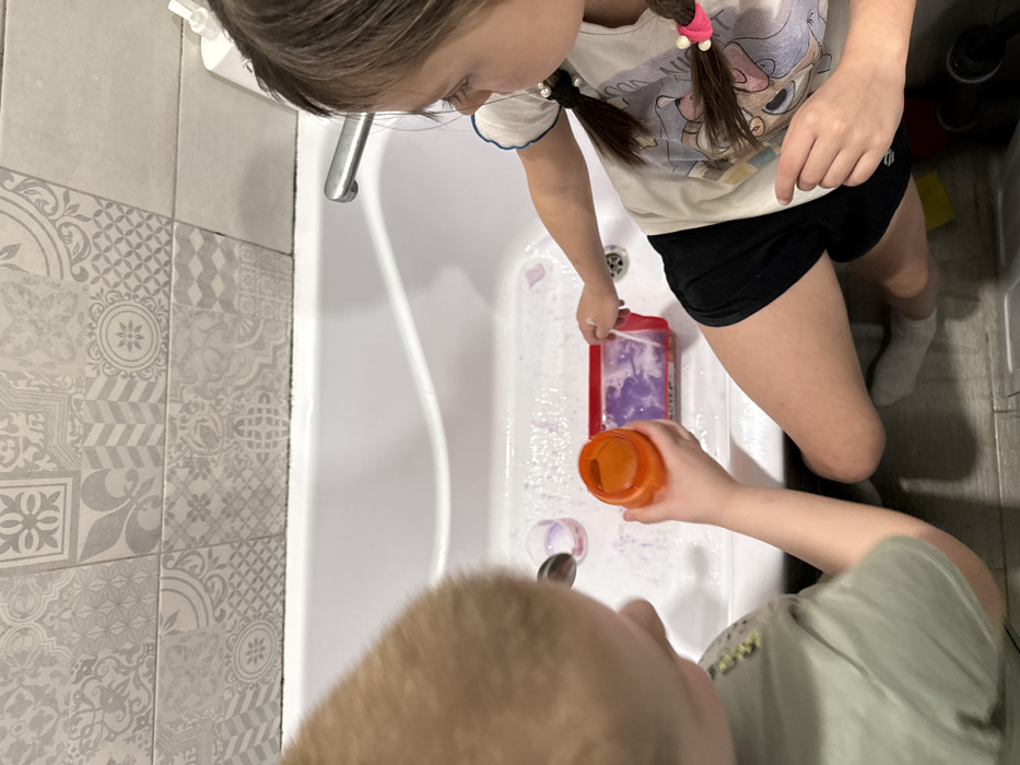 Two children stand over a white bathtub. One child, wearing black shorts and a cartoon t-shirt, holds a red container with purple liquid inside. They are looking down at the tub. The other child, wearing a green shirt, pours orange liquid from a container into the tub. A spoon lies inside the tub near a small amount of purple liquid. The bathroom has gray and white patterned tiles on the wall beside the tub.