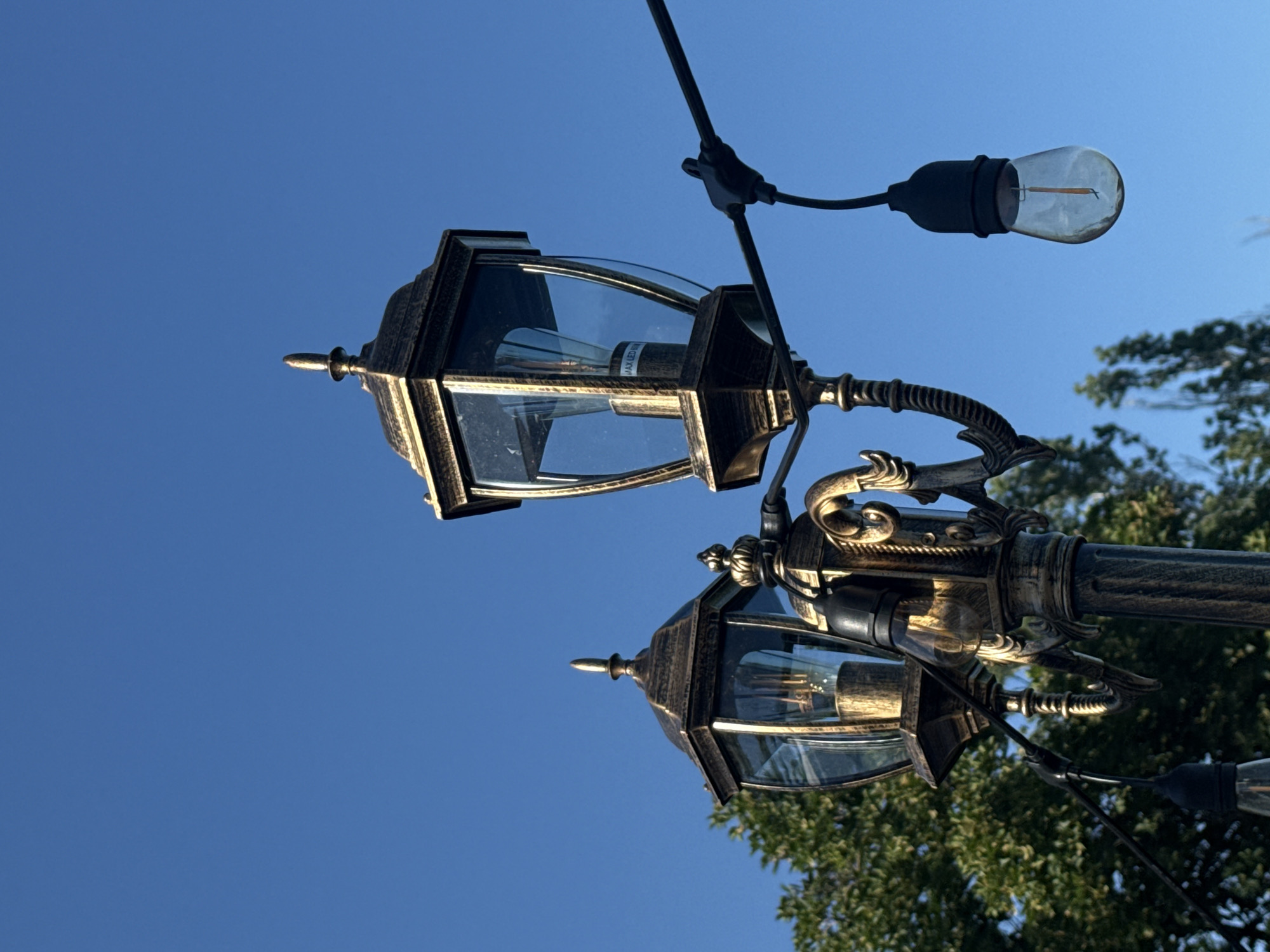 This is a close-up, low-angle shot of a decorative lamppost with a dark bronze finish, featuring ornate scrollwork and two distinct lanterns. The larger lantern is in the foreground, its glass panels revealing an Edison-style bulb. A second, smaller lantern is positioned to the left and slightly behind the first. A string of individual bare bulbs hangs from a black wire, with one bulb visible in the lower right quadrant of the image. The background is a clear, bright blue sky, with the upper branches of green trees visible on the left and right edges. The image appears to have been taken outdoors on a sunny day. Without further information, the specific location in Varna, Bulgaria cannot be determined from the image itself.
