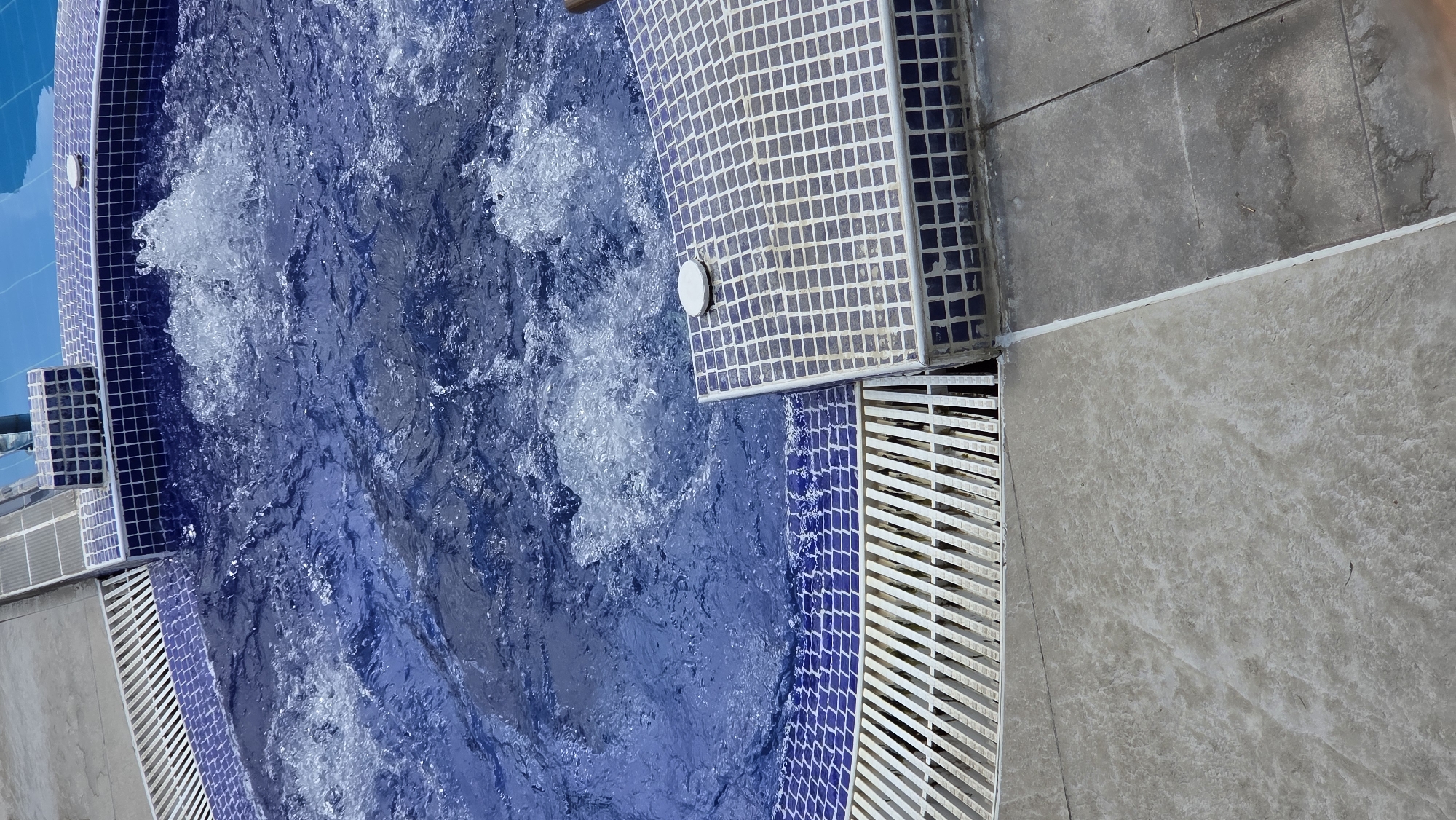 The image shows an overhead view of a section of a blue tiled swimming pool with bubbling water. The pool has a tiled edge with white and blue square tiles. There is a white drainage grate along the edge of the pool. The surrounding area is paved with grey and beige tiles. A small white circular object is visible on the tiled edge. The location is Unknown, Unknown.