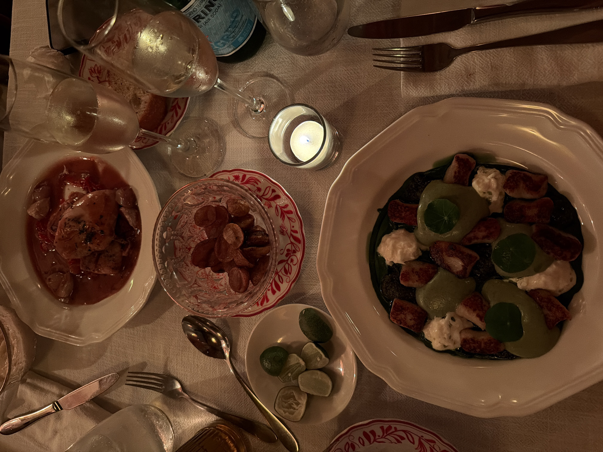 A high-angle shot shows a dimly lit table setting, likely in a restaurant, with various dishes and dining accessories. The location is not possible to determine based on the image alone; the prompt's mention of North Kuta, Indonesia, cannot be verified.
The table has a white tablecloth, likely linen, with subtle patterns. Visible are two white, scalloped-edged plates: One holds a red, gravy-like dish with what appears to be chicken or fish pieces, and the other contains a dish with dark green sauce, chunks of what looks like meat, dollops of white cream, and green herb garnishes.
There's a small, shallow white dish with several green limes or citrus wedges. A small glass bowl with a red and white patterned rim holds what appear to be small, round sausages or meat pieces.
Cutlery, including forks and knives with silver-colored handles, is arranged on the table near the plates. A silver spoon is near the small bowl of sausages.
A small, glass candle holder with a lit white candle provides some illumination. Two wine glasses, partially filled, and a bottle of sparkling water sit near the center. Another glass sits empty near the bottle.