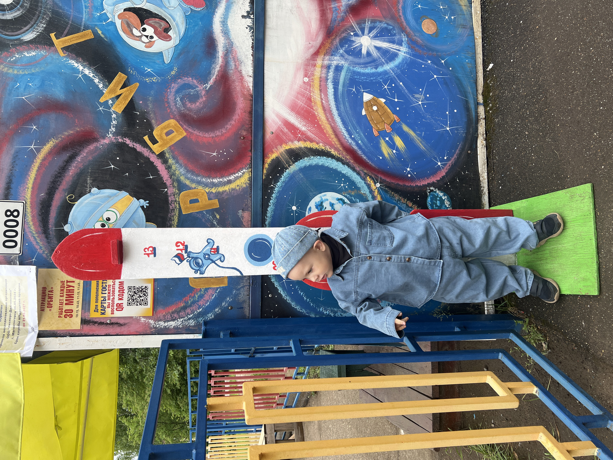 An outdoor scene in Chaikovsky, Russia, depicts a young child standing next to a rocket-shaped height measurement device. The child, dressed in a light-blue denim shirt, matching pants, and a denim beanie with a black undershirt, stands on a rectangular green wooden platform. The child's right hand rests on a blue metal railing composed of vertical yellow and horizontal blue bars. The child looks to the left of the frame.
Behind the child and the height measurement device, a vibrant, multi-colored mural covers a wall, depicting a cosmic theme with swirling nebulae, stars, planets, and cartoon characters including a blue alien-like creature and another character in a space helmet. Gold Cyrillic letters spelling "ОРБИТ" (ORBIT) are integrated into the mural. The white rocket-shaped height device features red markings and the numbers "13" and "12," with a cartoon figure below "12."
To the left of the rocket, attached to a support structure, are several informational signs in Russian. A yellow sign identifies the location as "АТТРАКЦИОН «ОРБИТА»" (ATTRACTION "ORBITA") and states it "РАБОТАЕТ КАЖДЫЕ 30 МИНУТ" (WORKS EVERY 30 MINUTES) with specific times. Below this, a white sign indicates "ДЛЯ ПОПОЛНЕНИЯ КАРТЫ ГОСТЯ" (TO REPLENISH GUEST CARD) using a QR code. An uppermost white sign references "г. Чайковский" (city Chaikovsky) and the extension of the attraction's service life until May 31, 2027. A yellow fabric awning is visible above the signs. A white plaque with the number "0008" is affixed higher on the wall.
The ground is a dark paved surface with some patches of dirt and sparse green vegetation. In the background, beyond the metal railing, green trees and foliage are visible under an overcast sky.